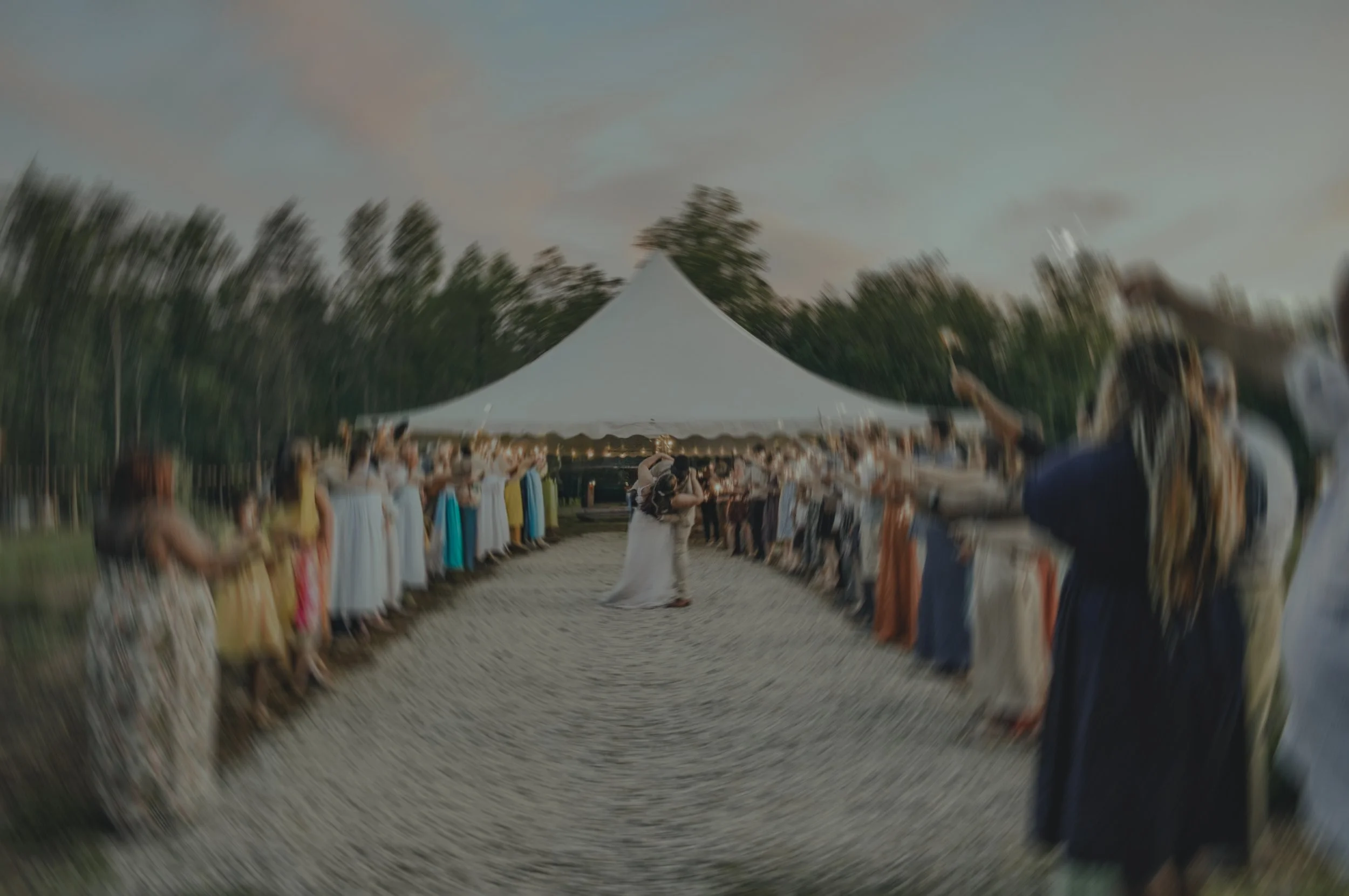 Couple kissing at outdoor wedding reception under a white tent, guests standing on either side, some with arms outstretched, in a natural setting with trees in the background, during dusk.
