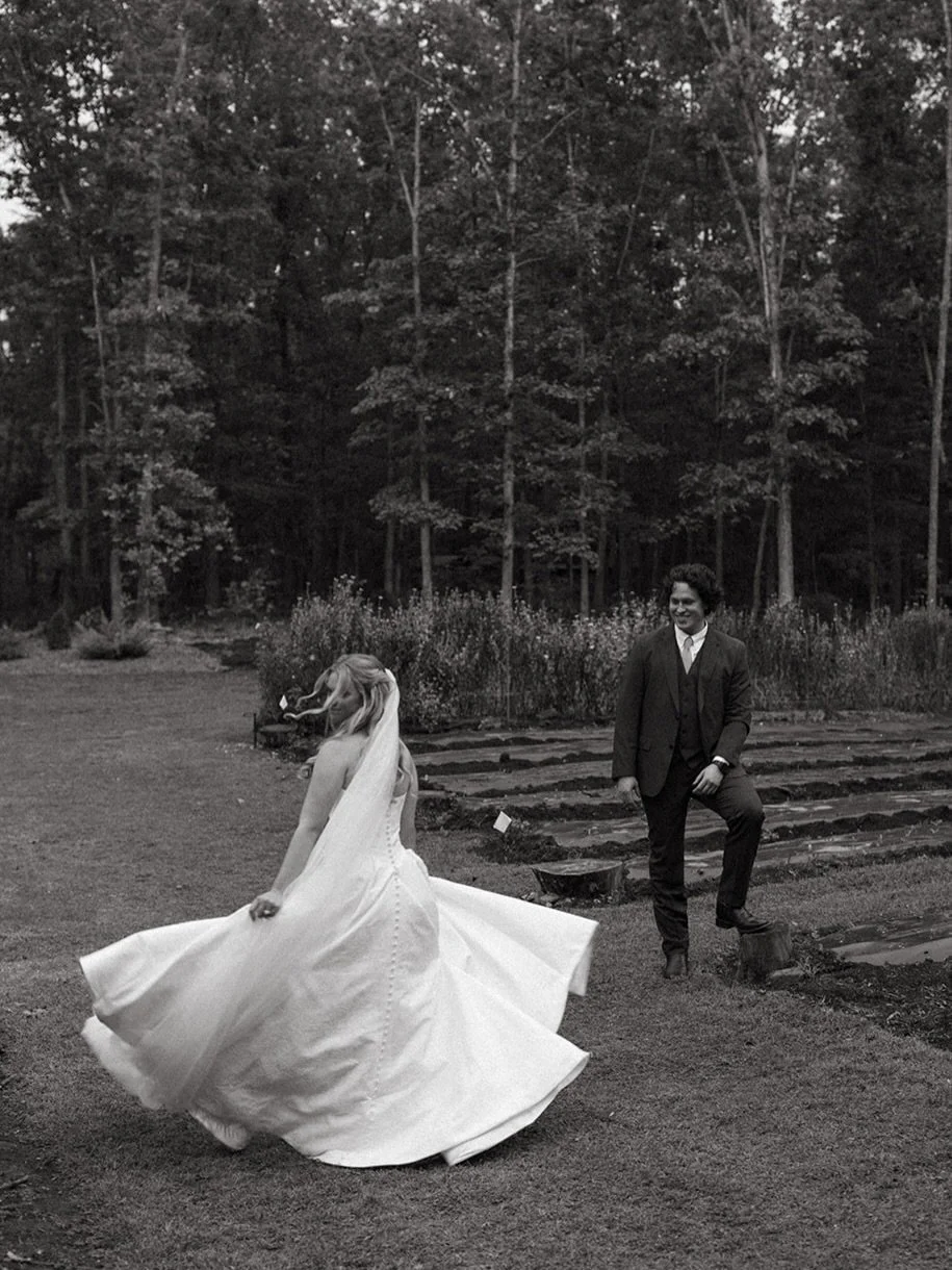A bride and a groom outdoors in a forested area, with the bride twirling in her wedding dress and the groom standing on a tree stump, smiling.