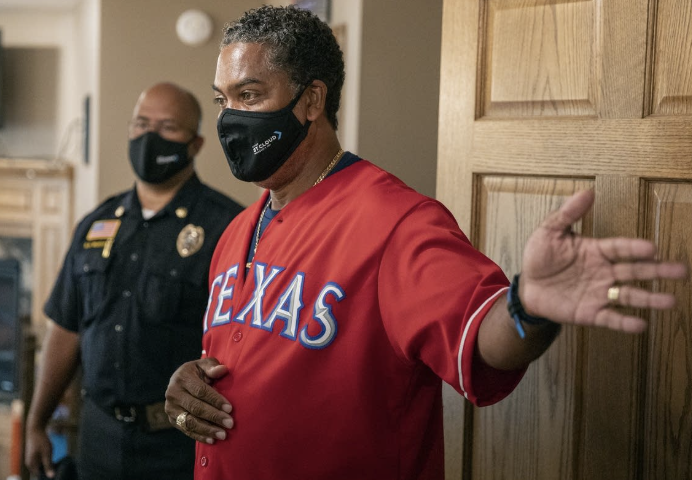 A man wearing a red Texas baseball jersey and a face mask gesturing with his right hand. Another man in a police uniform stands in the background.