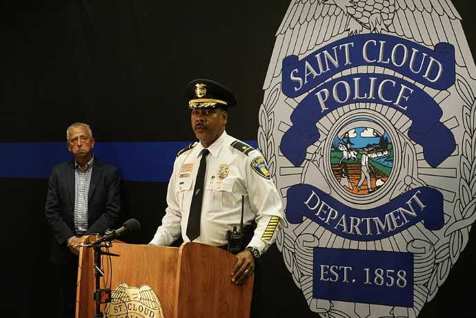 A police officer at a podium during a press conference, with a man standing behind him. The backdrop features the Saint Cloud Police Department logo.