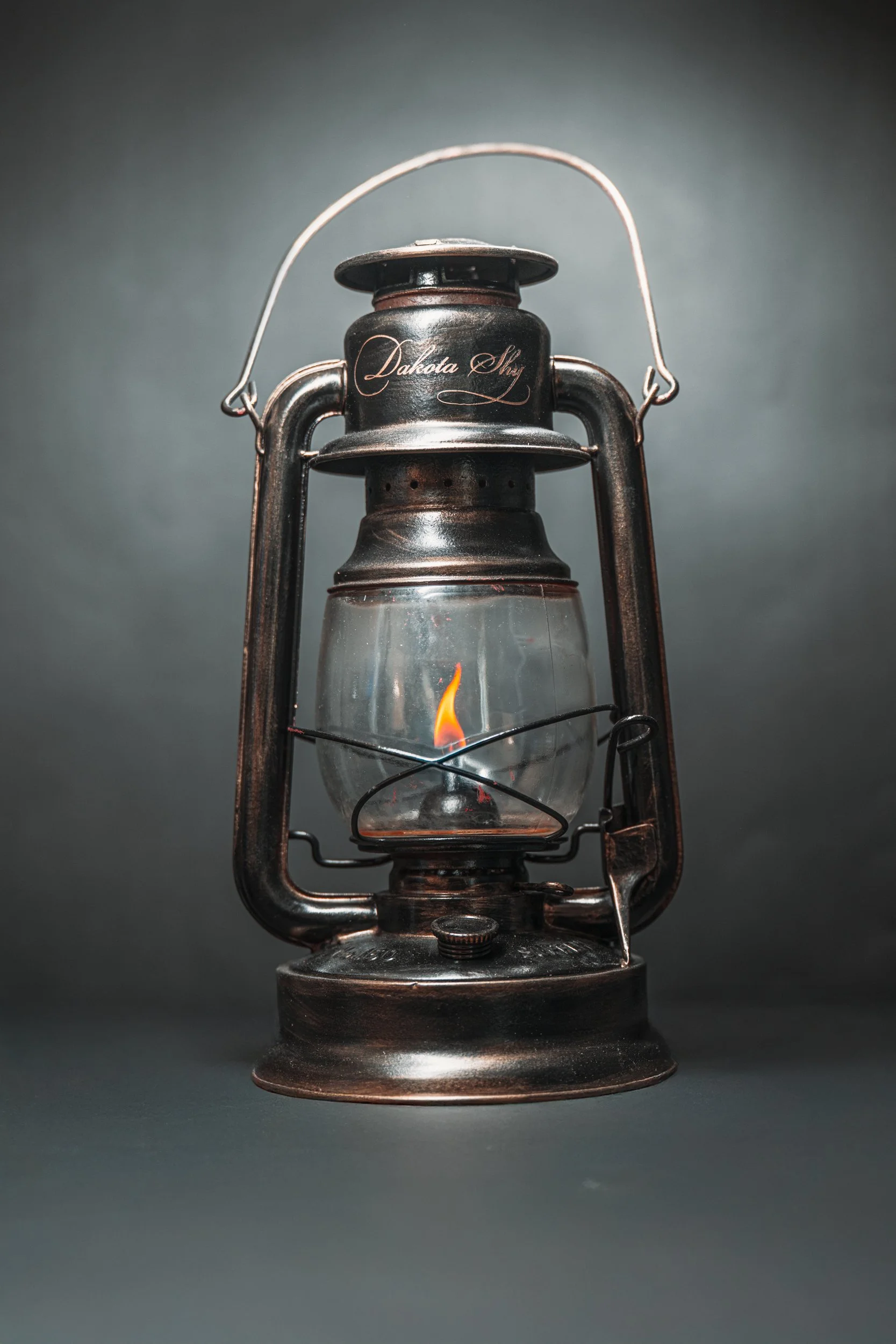 An antique oil lantern with a small flame inside, labeled Dakota Sky, on a neutral background.