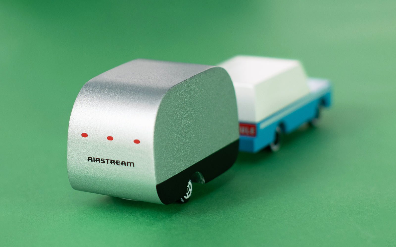 The back passenger side view of a silver and black painted, wooden airstream camper attached to a light blue car.