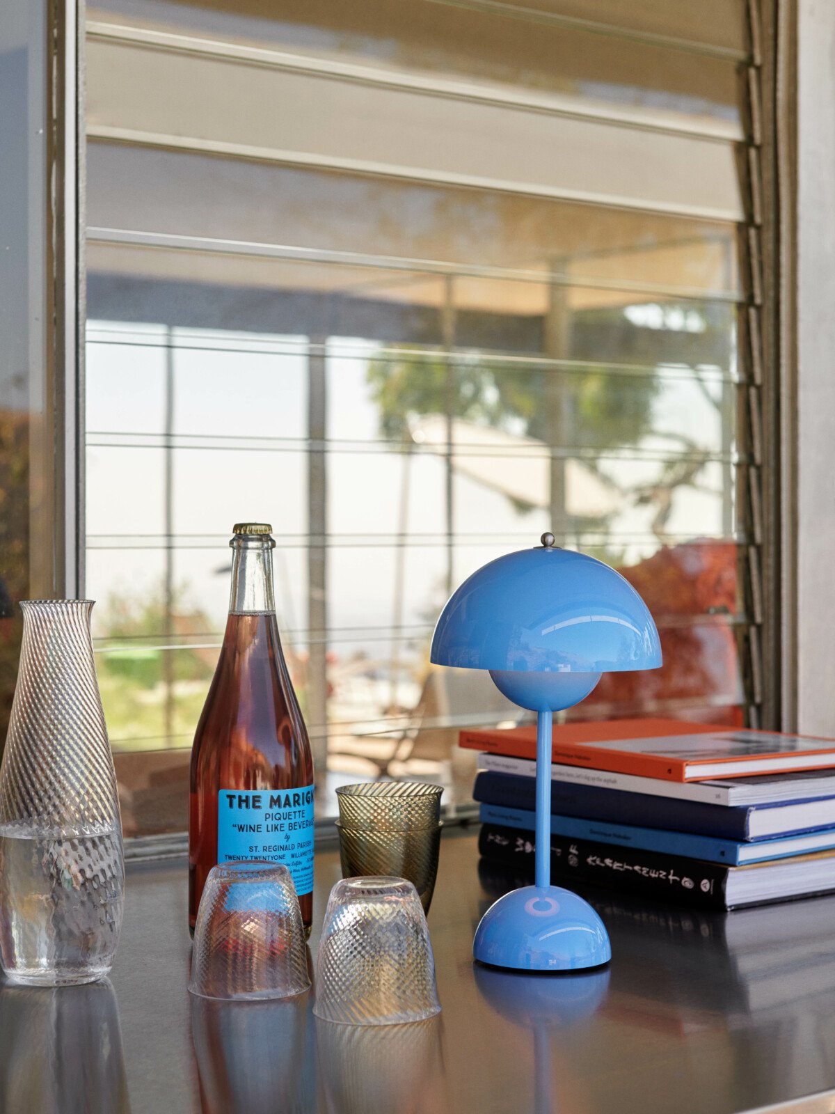 Swim Blue Flowerpot Lamp on a bar counter next to a glass carafe, a beverage bottle, and a stack of books.