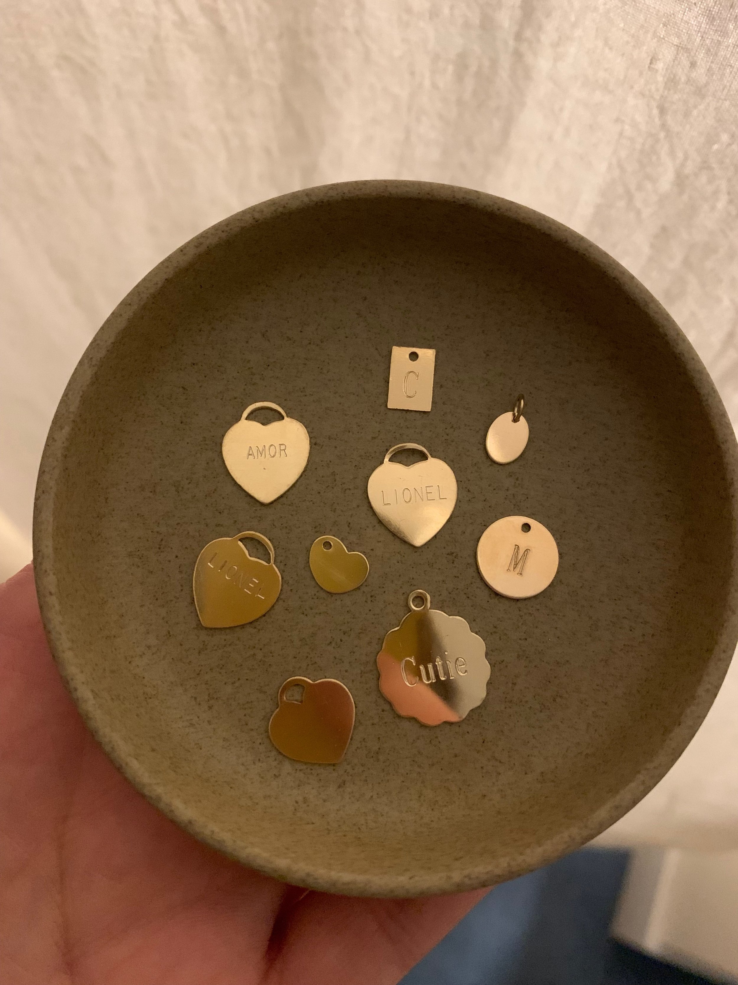 Various shapes of gold fill charms, including hearts, circles, and rectangles, on a brown ceramic dish, held in someone's hand. a