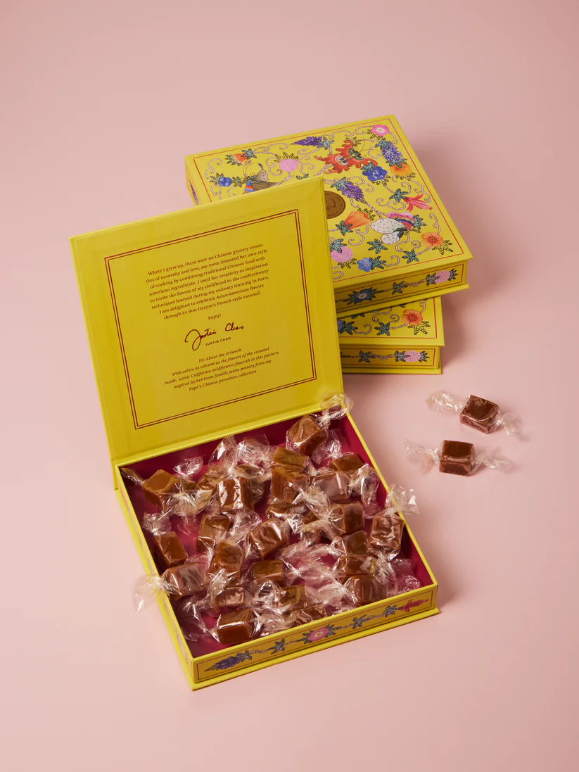An open box of Le Bon Garcon P.O.G. Caramels filled with individually wrapped caramels, next to two wrapped caramels outside of the box and two stacked, closed boxes of caramels.