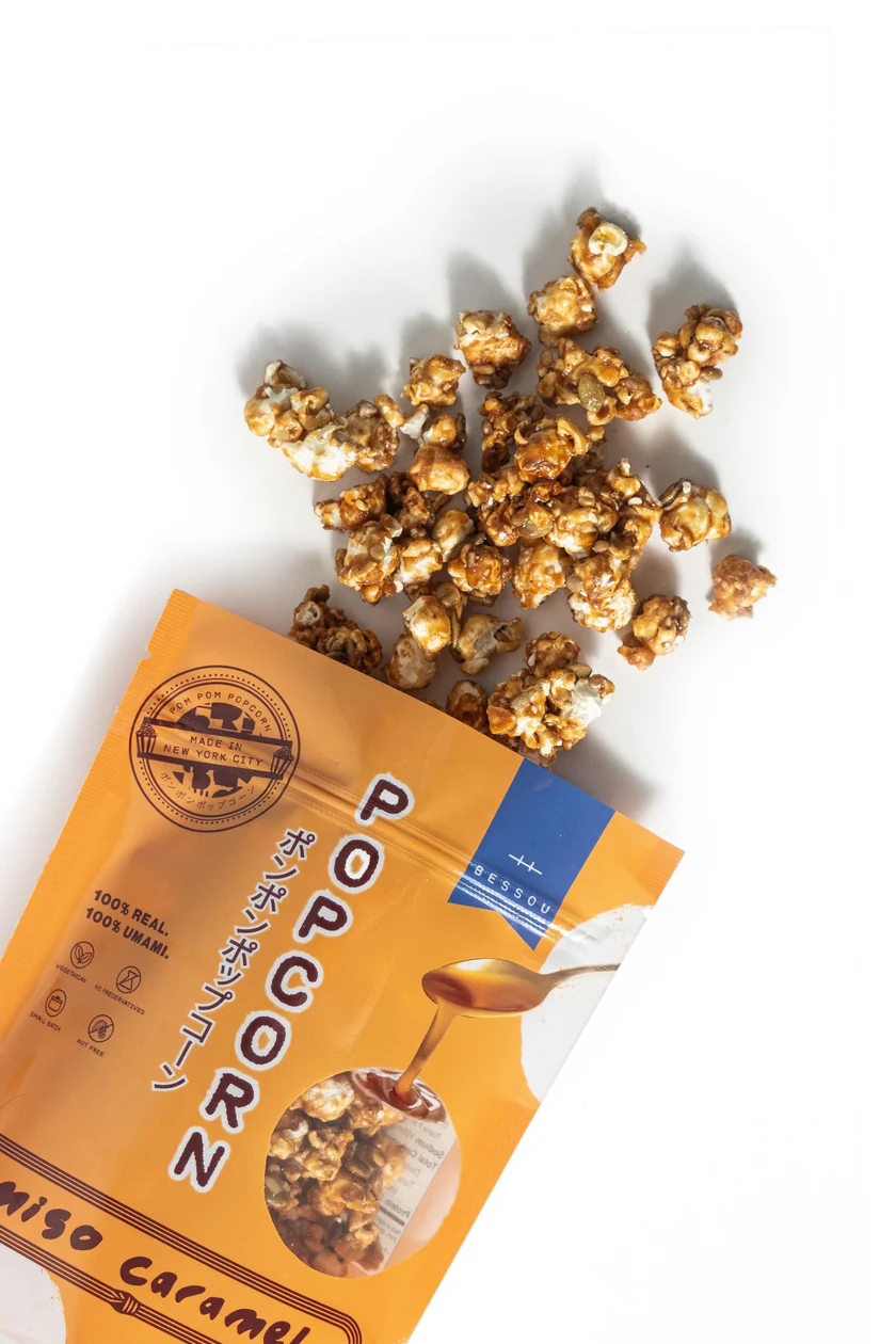 An open, orange package of Bessou Miso Caramel Popcorn spilling onto a white surface.
