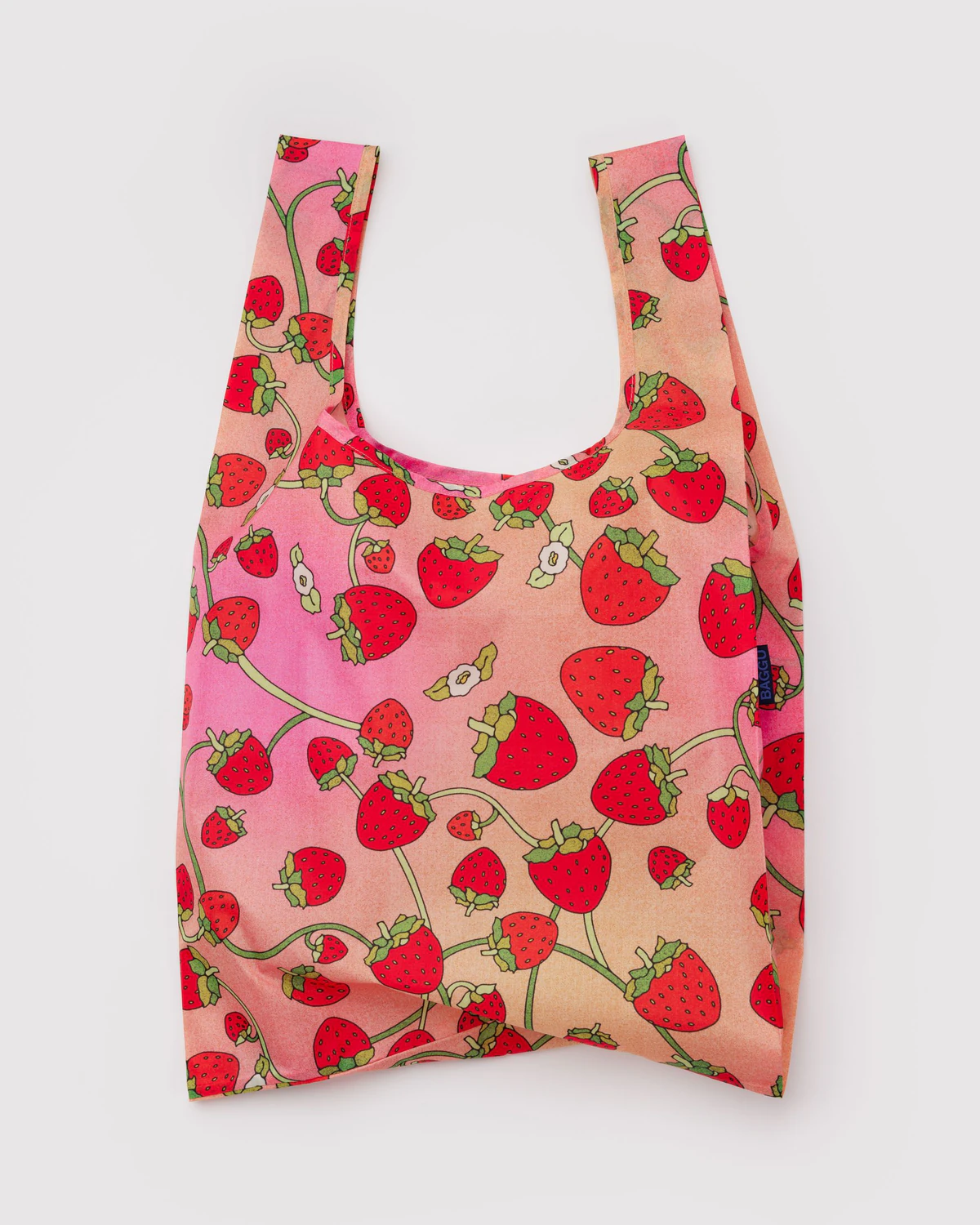 Strawberry Blossom Standard Baggu, a reuseable nylon tote with a pattern of strawberries on a vine on a peach and pink ombre background.