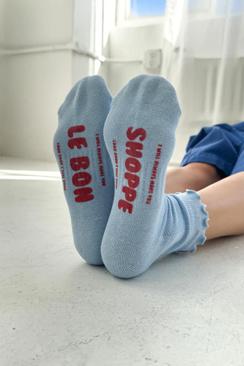 The feet of a model wearing the Ruffled Hugger Crew Socks in Periwinkle, with red bubbled words on the soles.