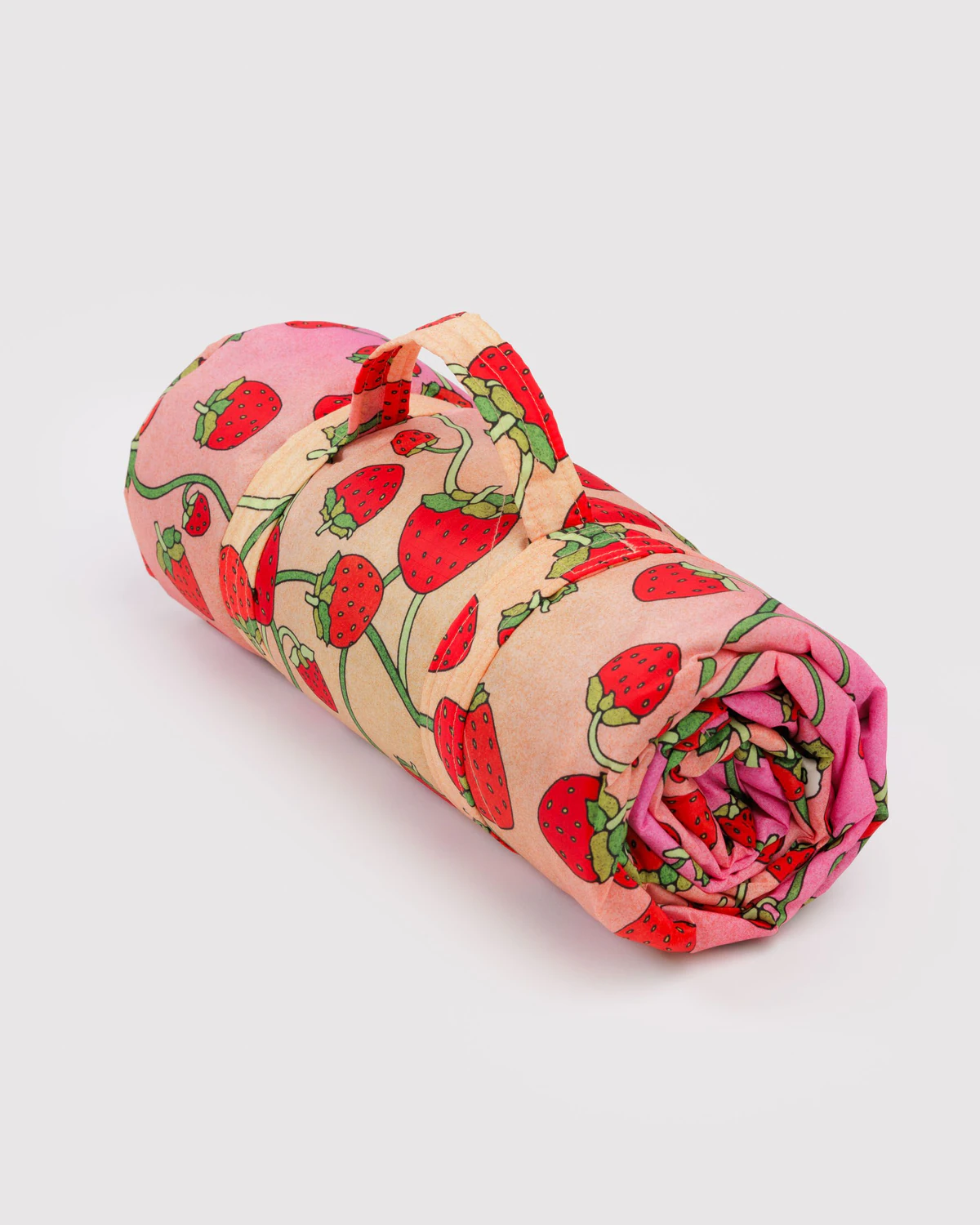 A rolled up Baggu Strawberry Blossom Puffy Picnic Blanket with a strawberry floral print on a peach  and pink ombre background.