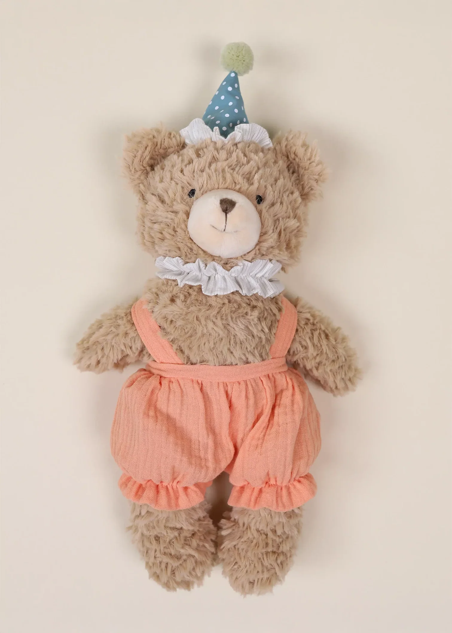 A plush, light brown bear wearing peach overalls and a blue party hat.