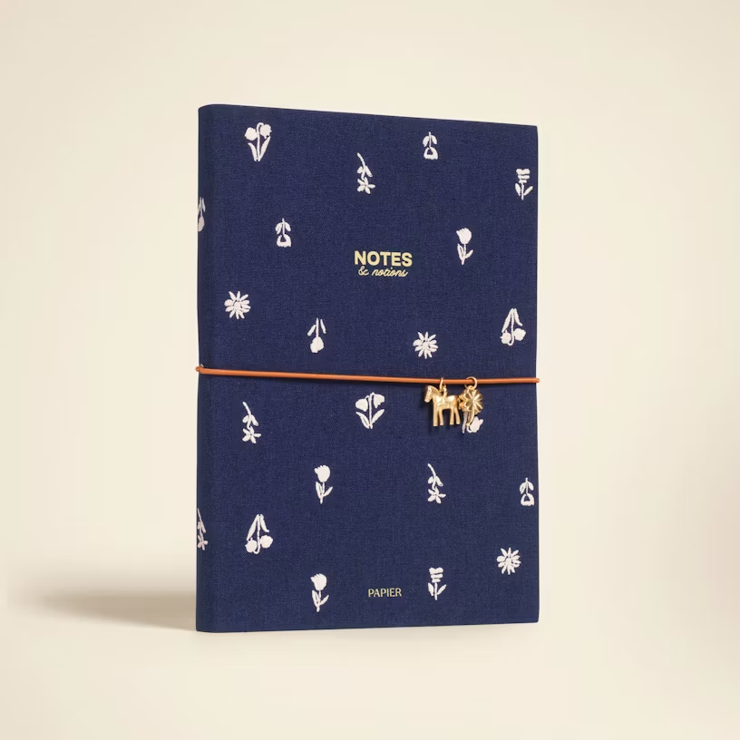 Papier Twilight Blooms Notebook, a notebook with a navy blue fabric cover with embroidered white flowers and an elastic closure with a gold horse charm and a gold clover charm.