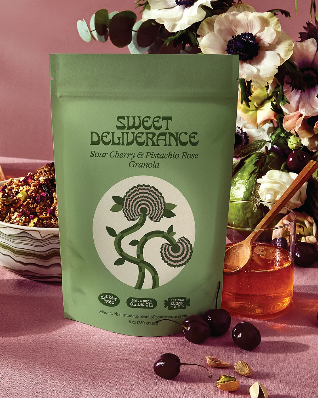 A green package of Sweet Deliverance Sour Cherry & Pistachio Rose Granola, with ingredients next to and behind it, including cherries and honey.