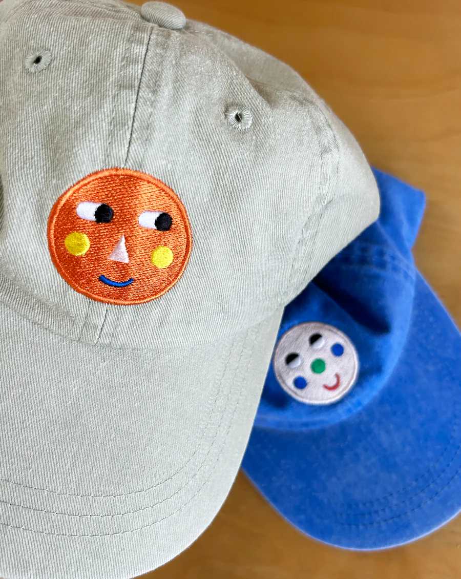 A khaki Feelings Cap with an orange embroidered face on top of a blue Feelings Cap with a light pink embroidered face.