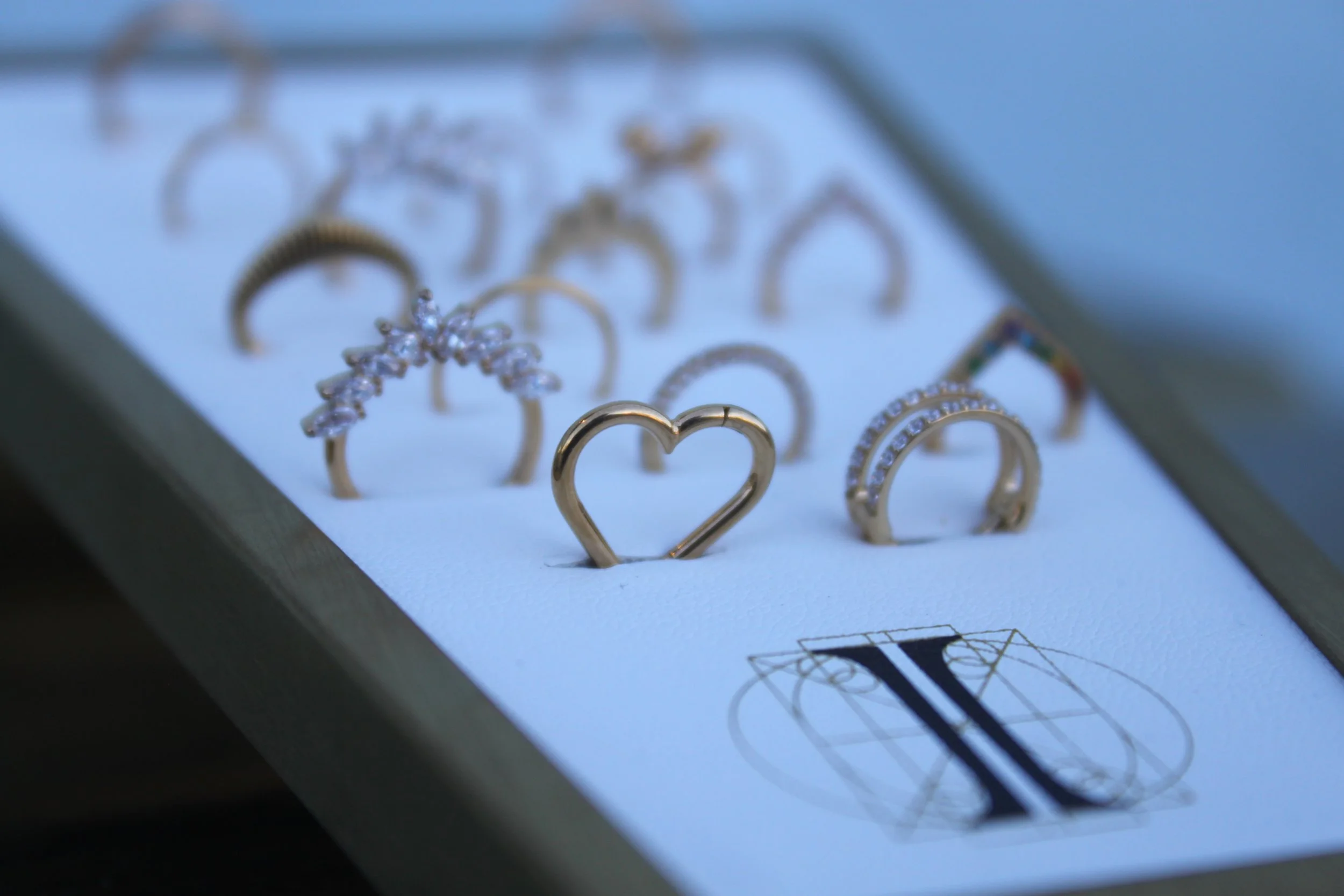 Close-up of a jewelry display box with gold rings, some adorned with small gemstones, arranged on a white surface. The display is partially out of focus.