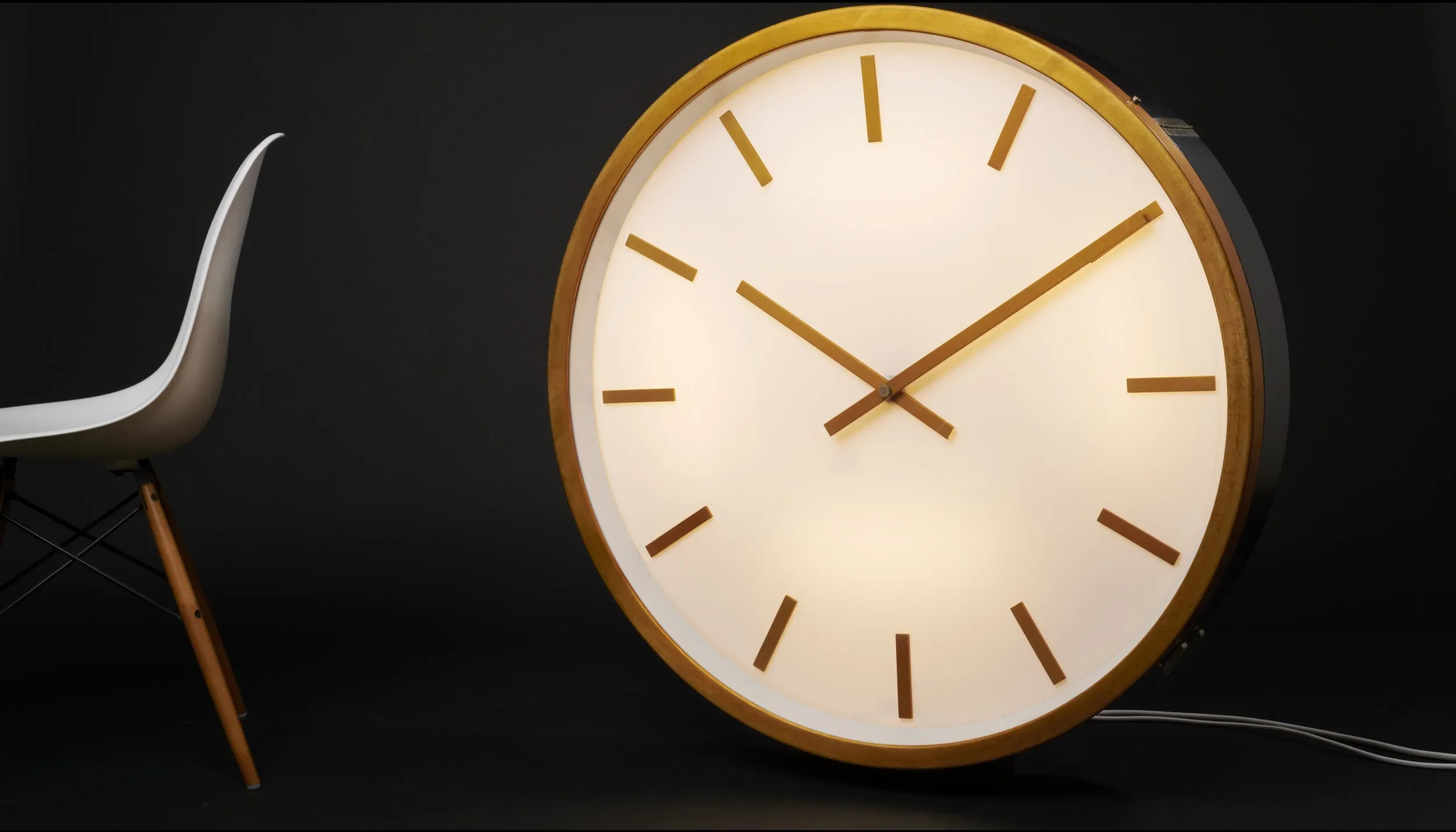 A round wall clock with a wooden frame and backlit white face, showing the time as 2:12. To the left, a modern white chair with wooden legs is partially visible against a black background.