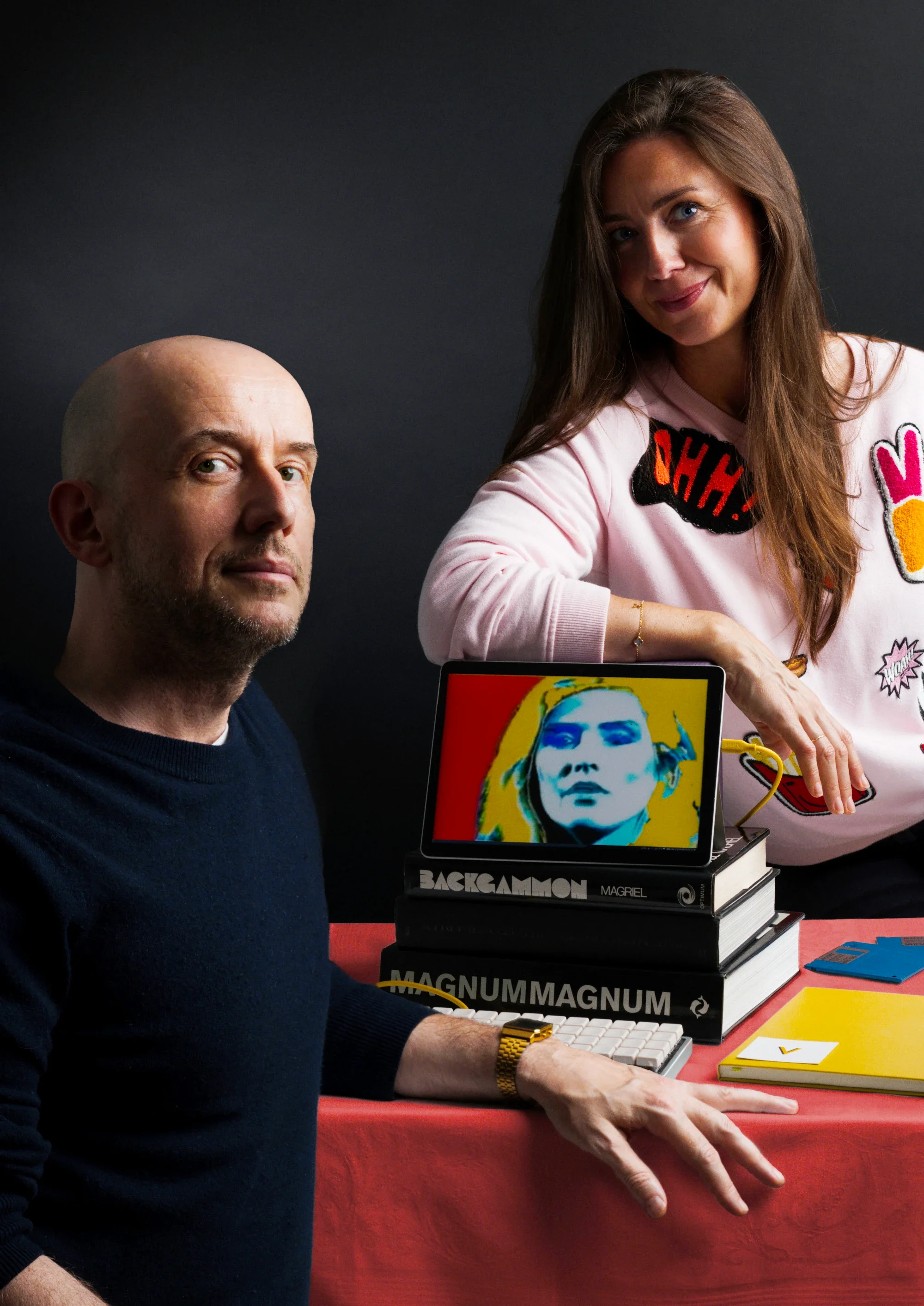 Vanessa and Martin at a red-covered table with a tablet showing a pop-art portrait of Debbie Harry, a keyboard, art books and floppy disks.