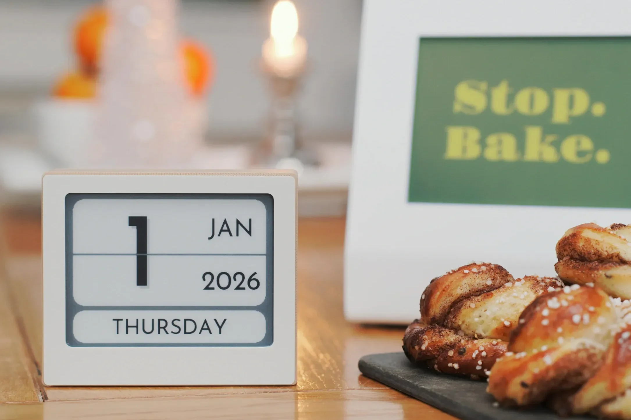 A desk calendar showing Thursday, January 1, 2026, placed beside a plate of cinnamon rolls, with a sign in the background that reads 'Stop. Bake.'