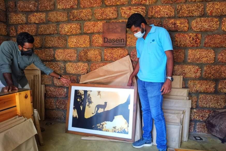 Two men wearing face masks handling a framed silhouette art piece of a leopard on a tree branch inside a room with a brick wall.