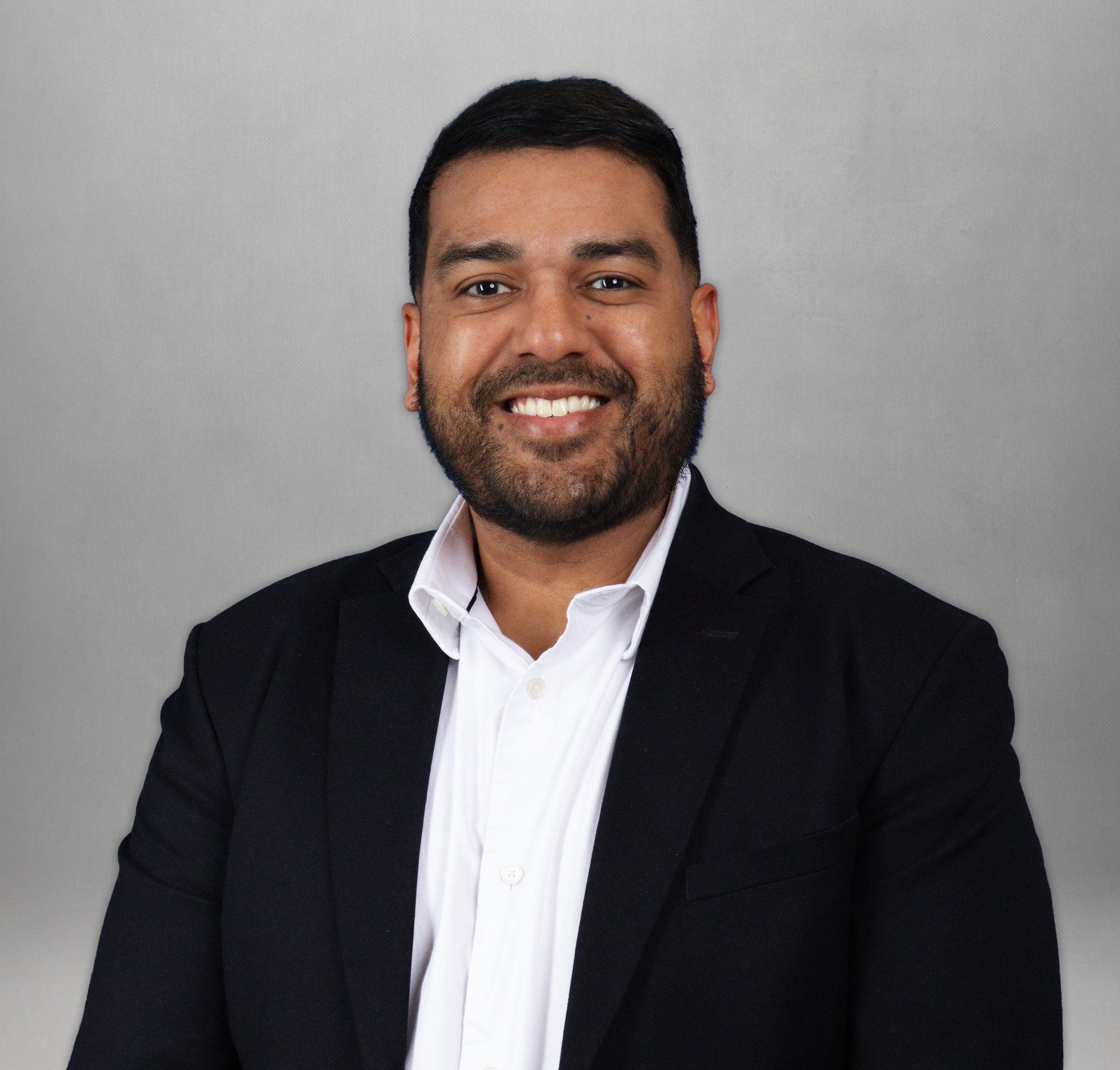 A man with a beard and mustache dressed in a dark suit, white shirt, and light blue striped tie, smiling in an indoor setting.