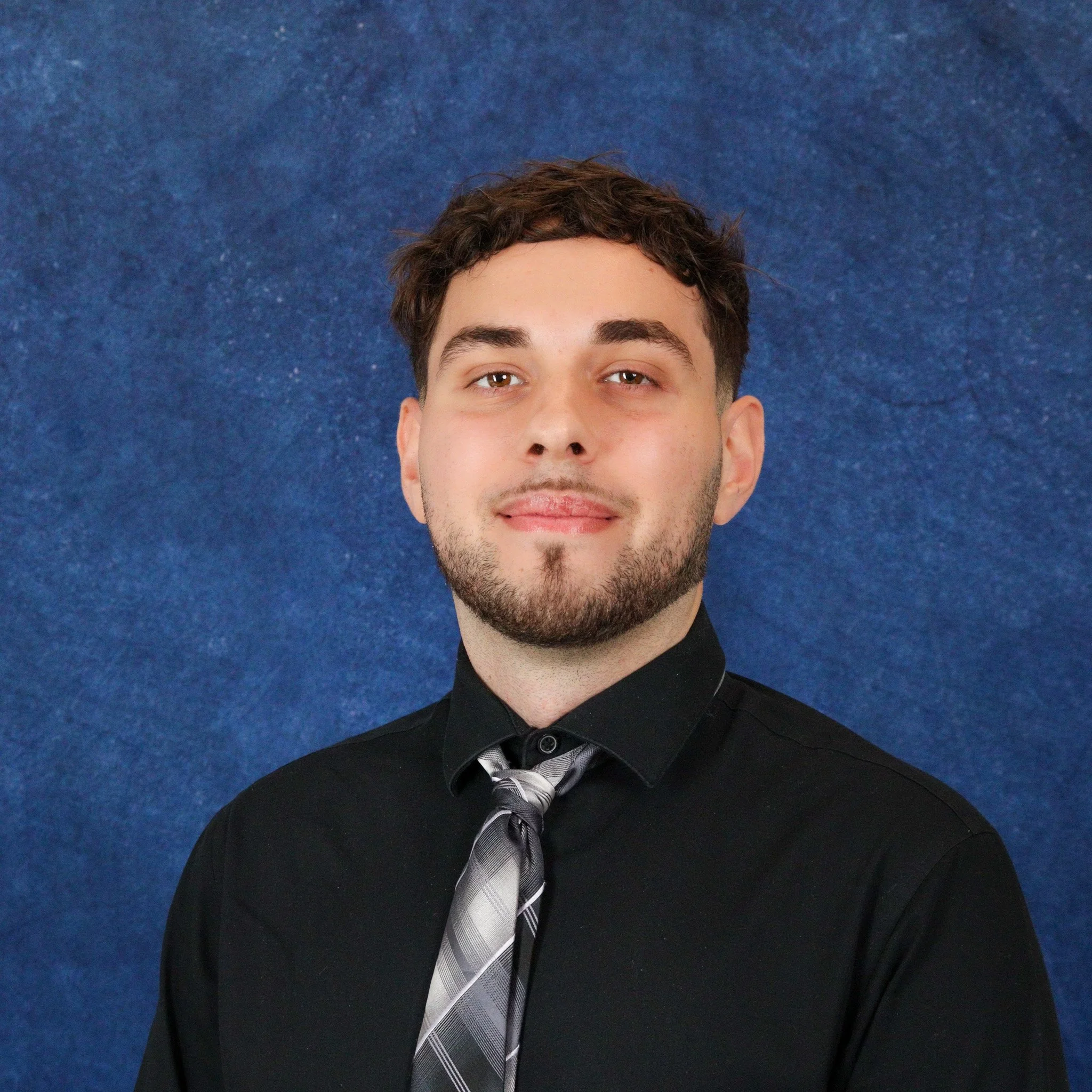 A young man with short brown hair and a beard, wearing a black shirt and a gray plaid tie, smiles slightly in front of a textured blue background.
