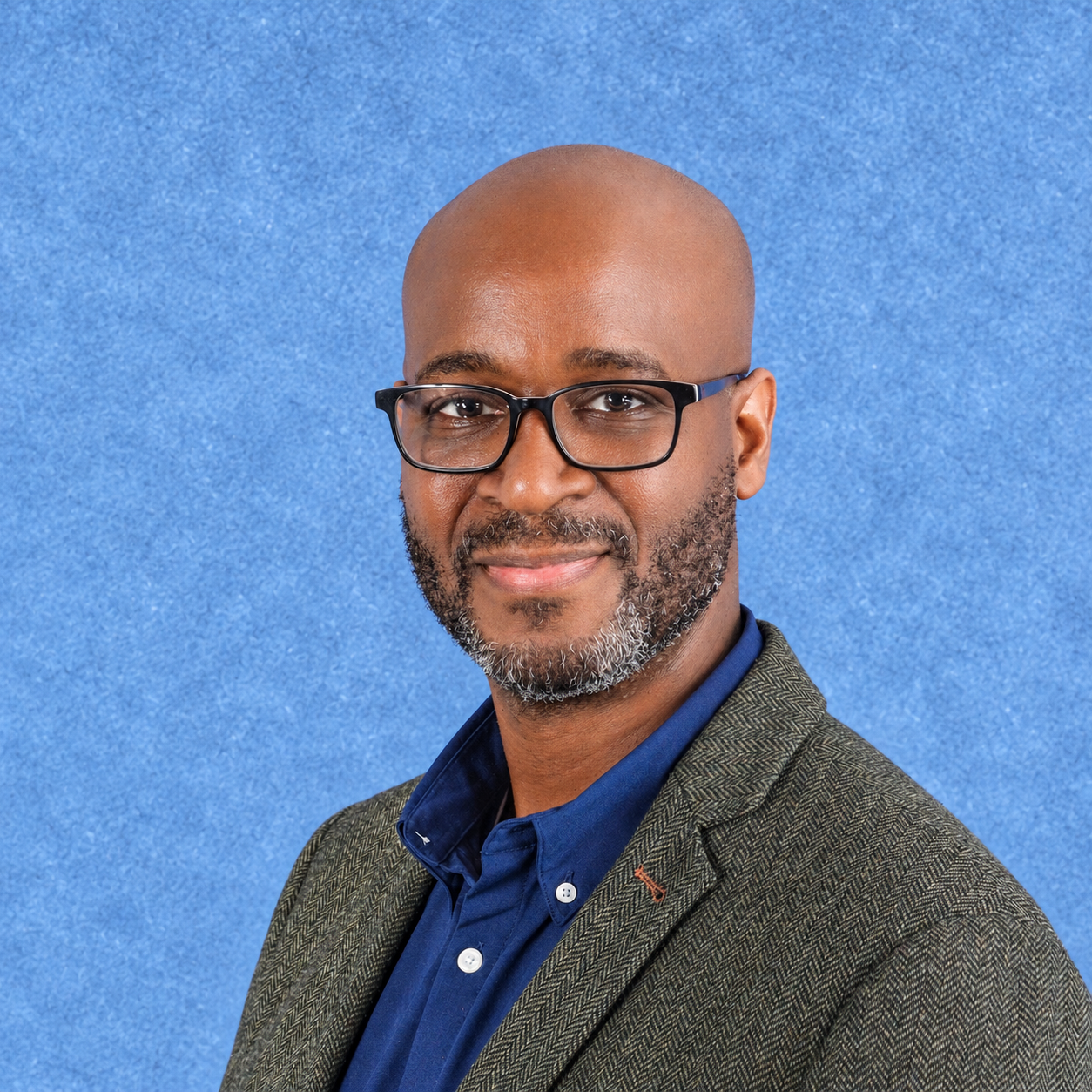 A middle-aged Black man with a beard and glasses, wearing a brown blazer and a striped shirt, smiling in front of a blue textured background.