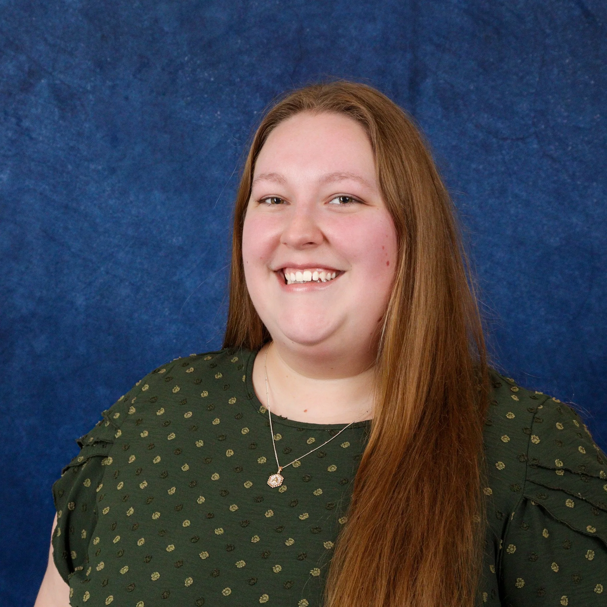 A young woman with long red hair, smiling, wearing a dark green blouse with a pattern of small gold circles, a silver necklace with a small pendant, standing against a blue textured background.