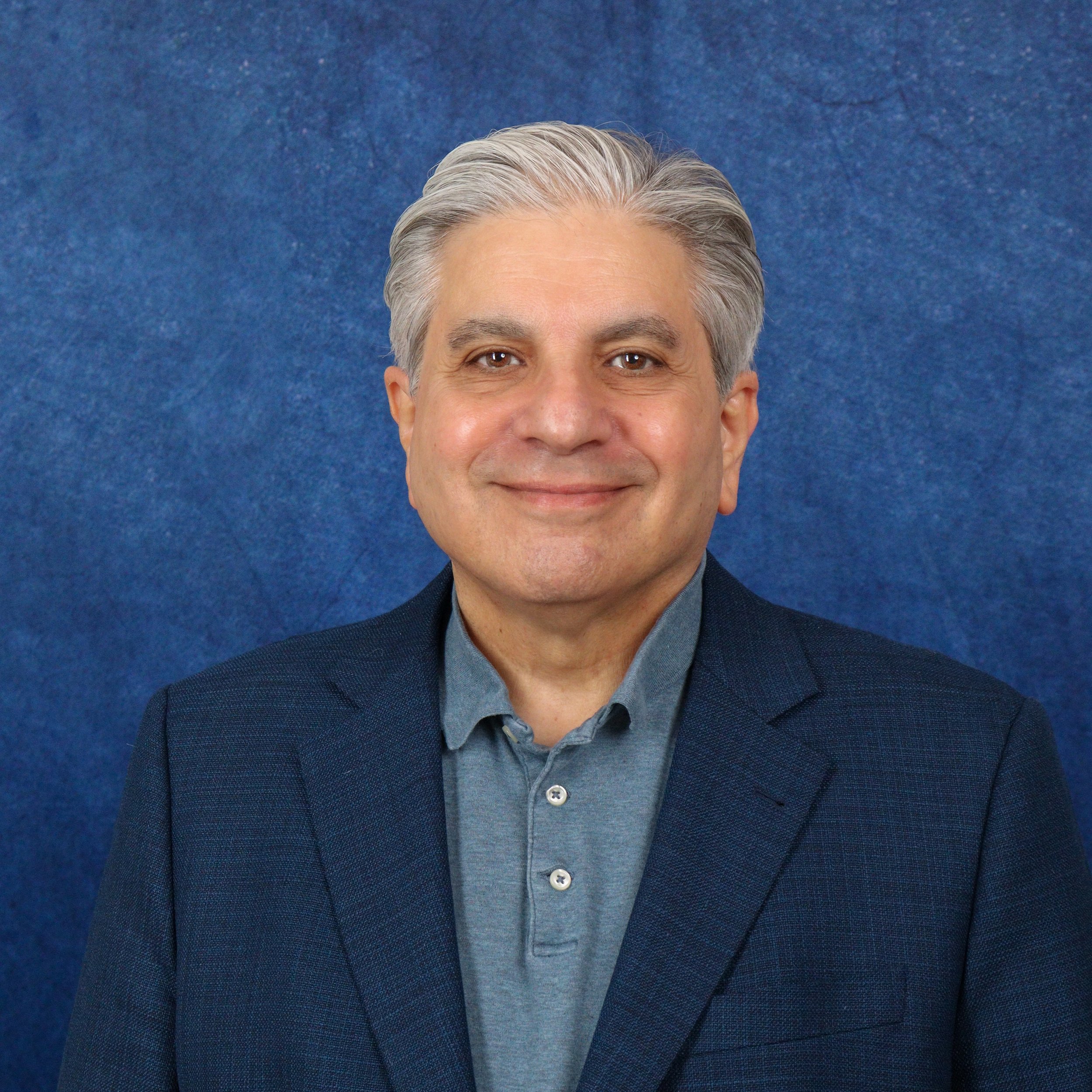 A middle-aged man with gray hair and beard, wearing a dark suit, light purple dress shirt, and red patterned tie, standing in front of a blue textured background.
