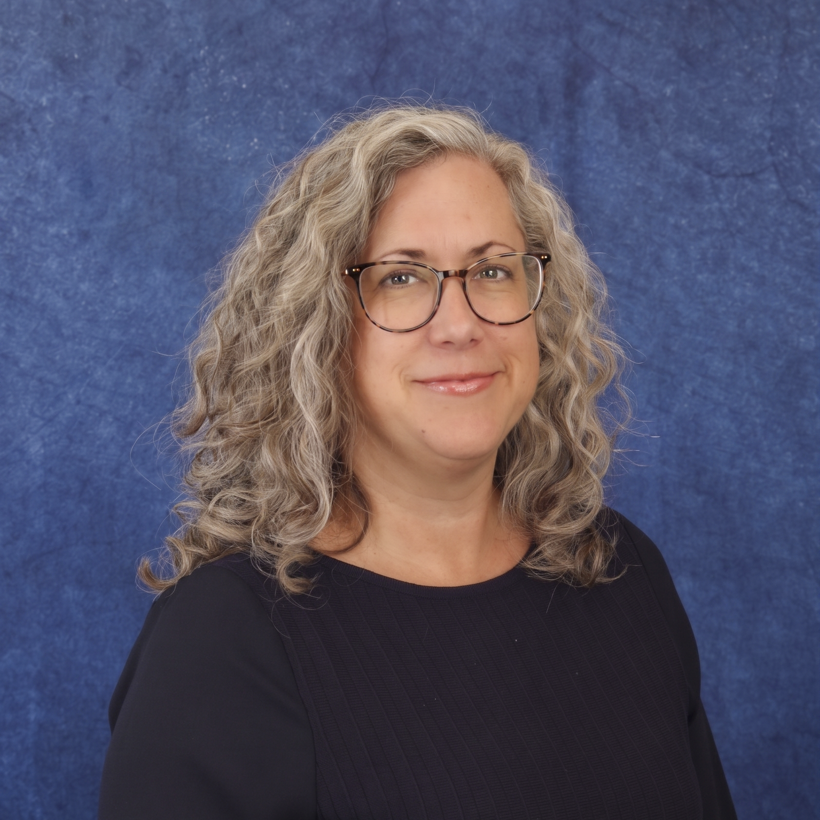 A woman with curly gray hair and glasses wearing a black blazer and green top, standing against a textured blue background.