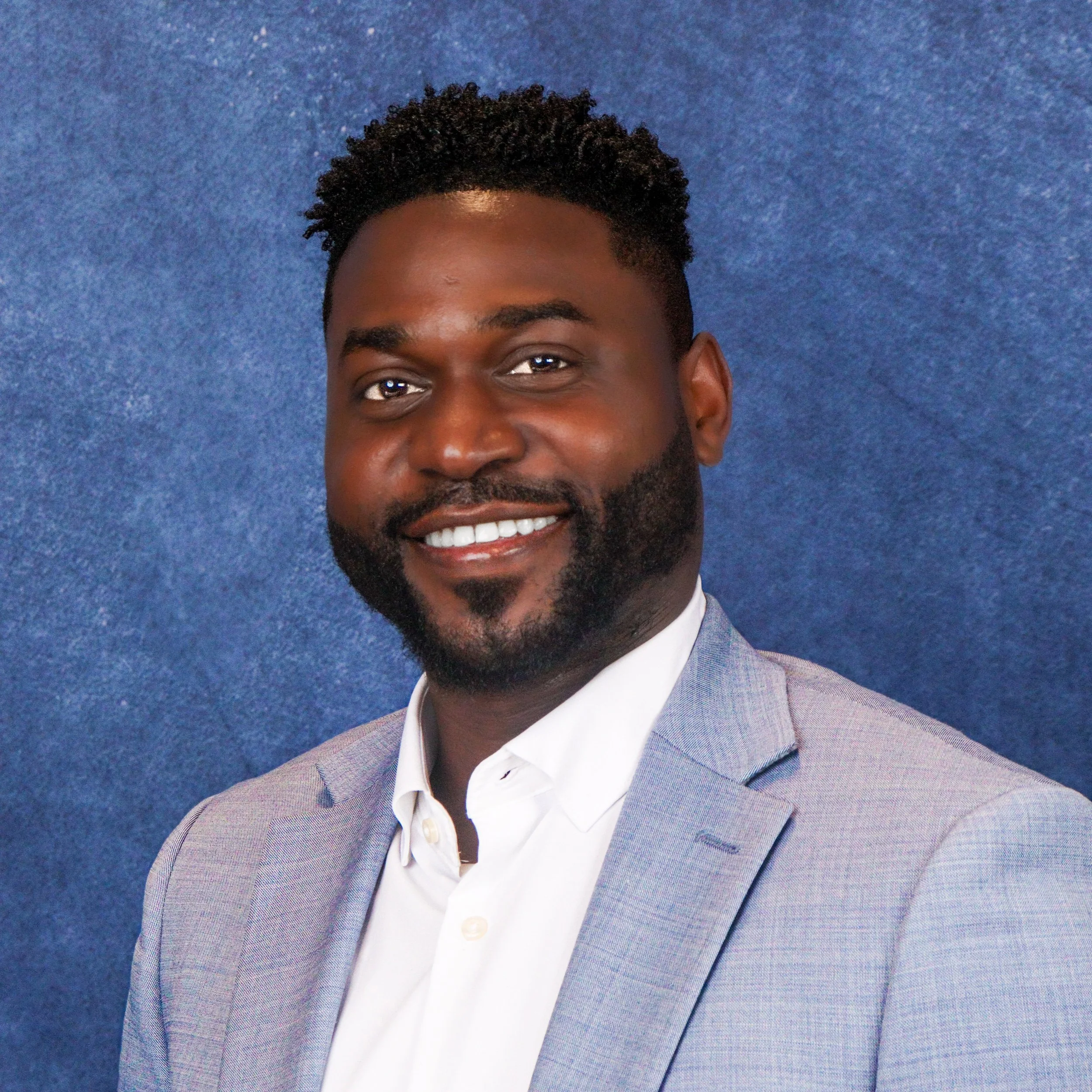 Professional man smiling in light gray suit jacket and white shirt against blue background.