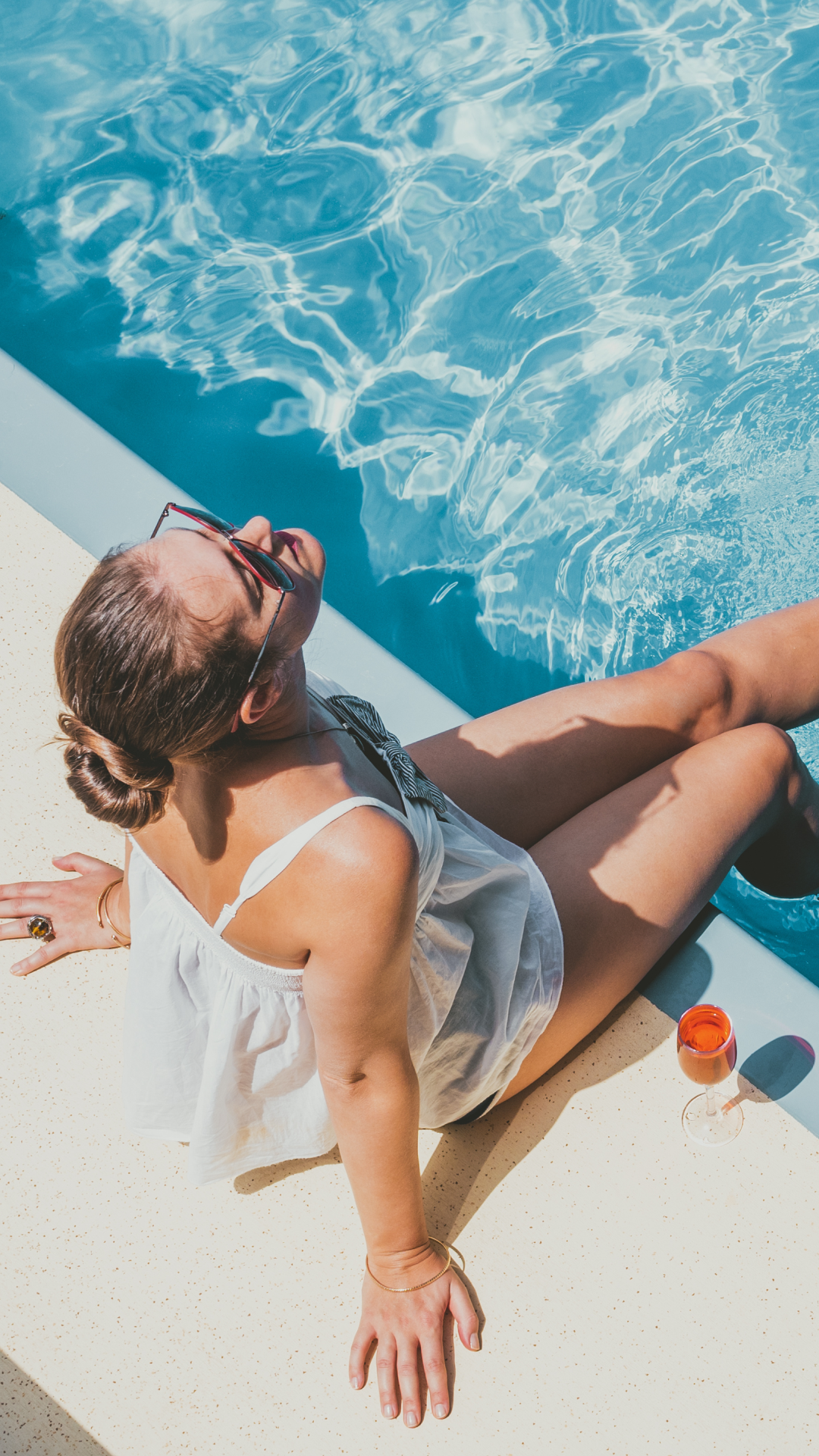 Woman in a white top relaxes by a pool, sitting on the edge with feet in water, wearing sunglasses. A drink is nearby, conveying a serene, sunny vibe.
