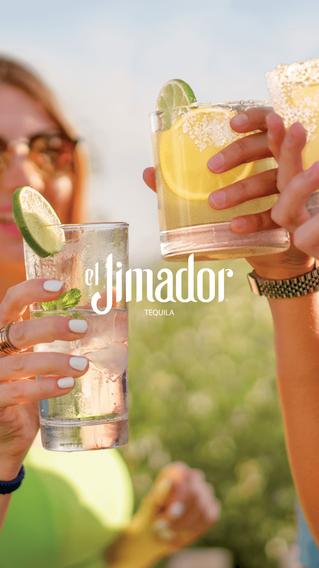 Two people clink glasses of cocktails garnished with lime slices and salted rims against a sunny, outdoor backdrop.