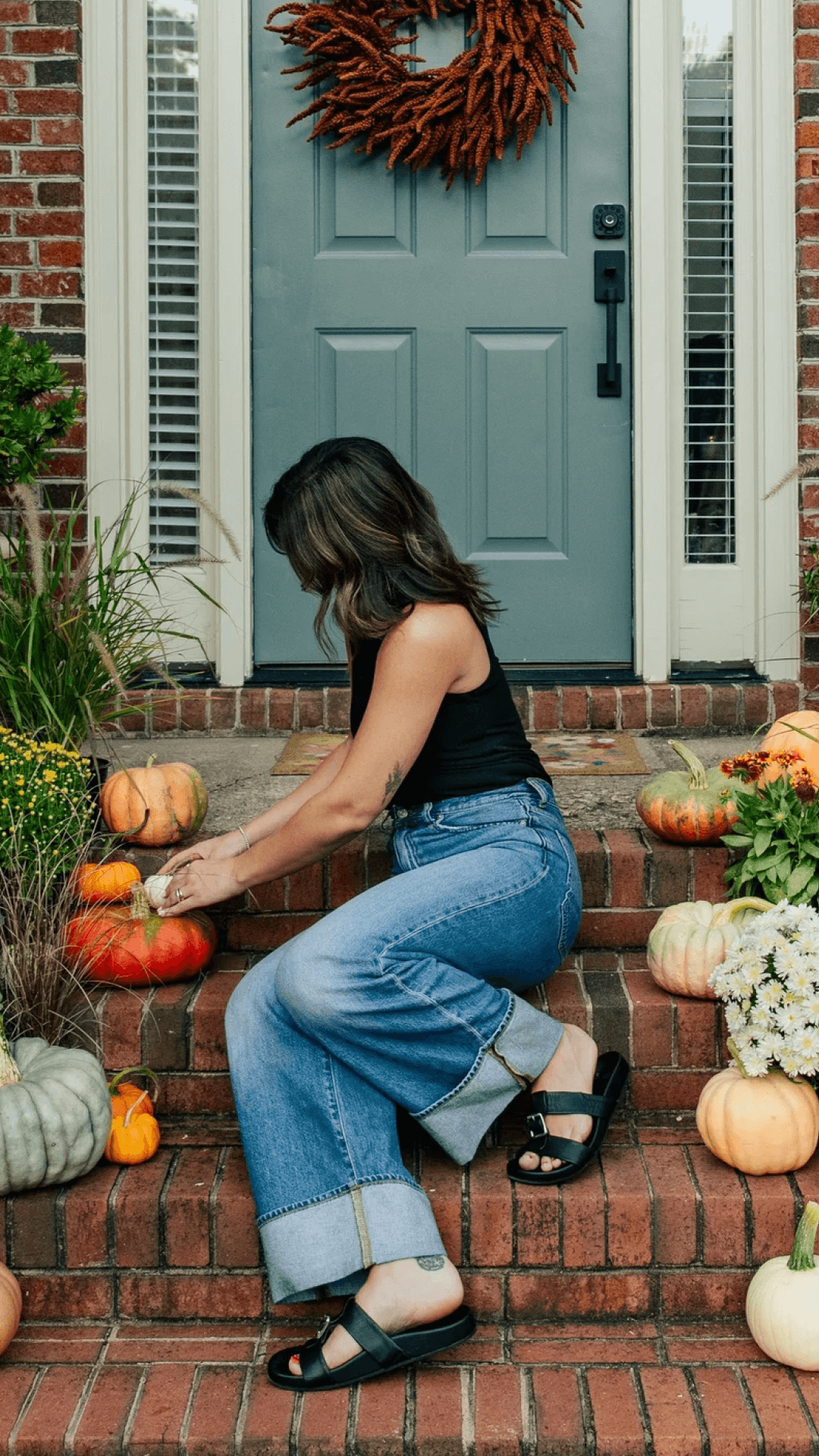 Woman in black tank and jeans arranging pumpkins on brick steps adorned with assorted pumpkins, greenery and fall wreath on the blue front door.