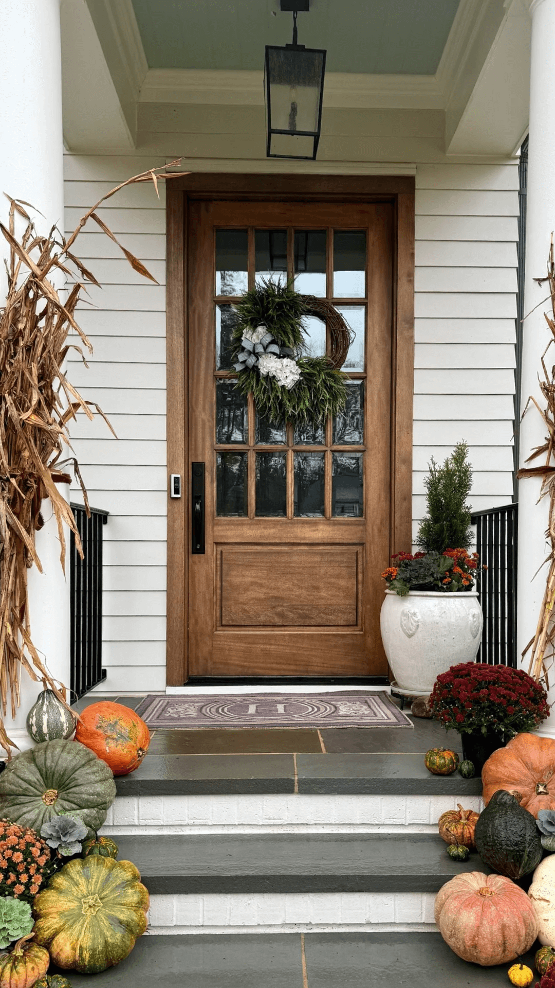 A welcoming front porch with a wooden door decorated with a wreath and surrounded by pumpkins and autumn flowers, creating a cozy fall atmosphere.