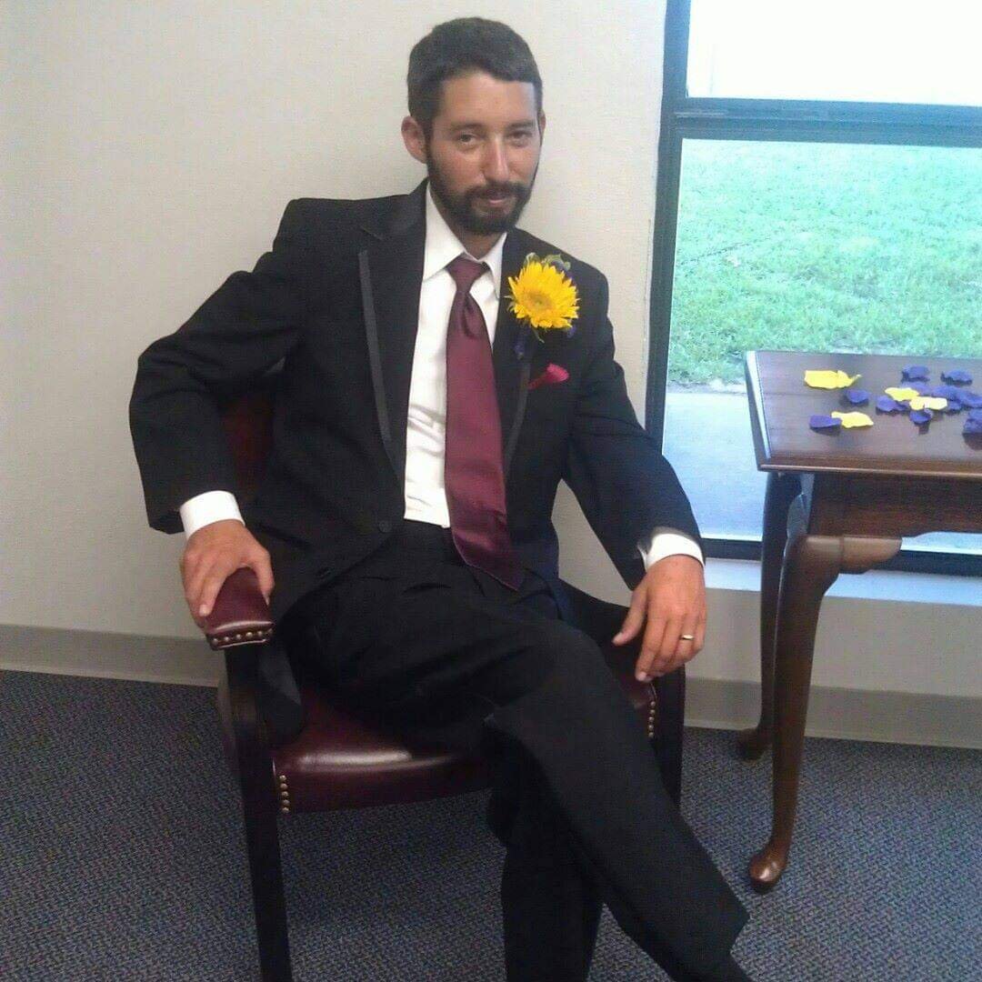 David is wearing a black tux with a white shirt and maroon tie. He has a yellow carnation pinned to the label. He is sitting cross-legged in a wooden chair next to a wooden table with yellow and purple confetti. He has a slight grin on his face.