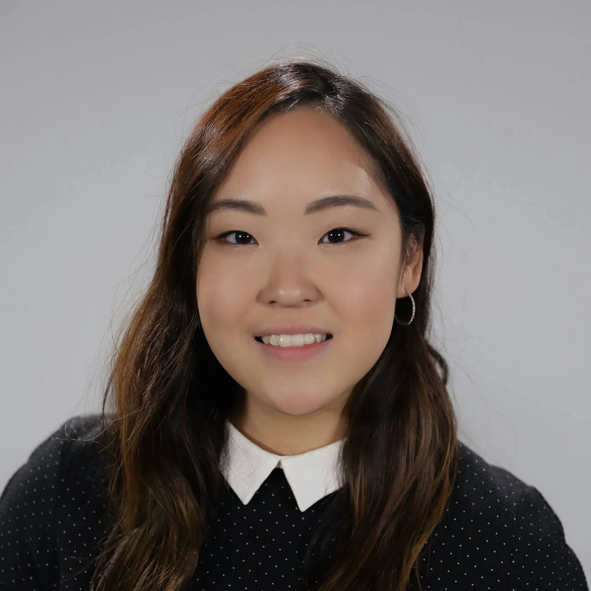 Portrait of a young woman with long brown hair, wearing a black polka dot top with a white collar, smiling against a gray background.