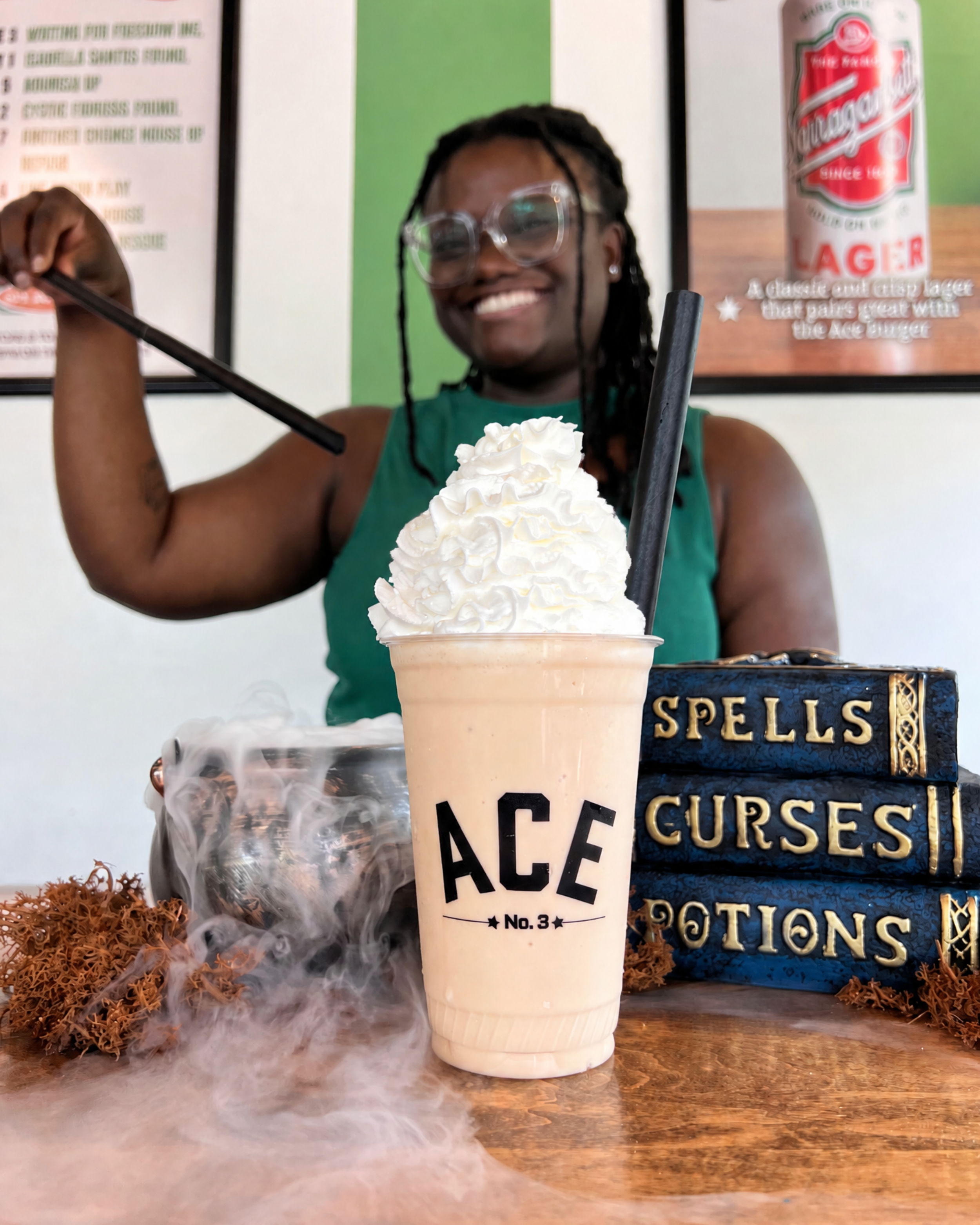 A smiling woman with glasses holds a magic wand in front of a milkshake with whipped cream in a restaurant. There is a decorative box labeled "SPELLS and CURSES" and a menu in the background.