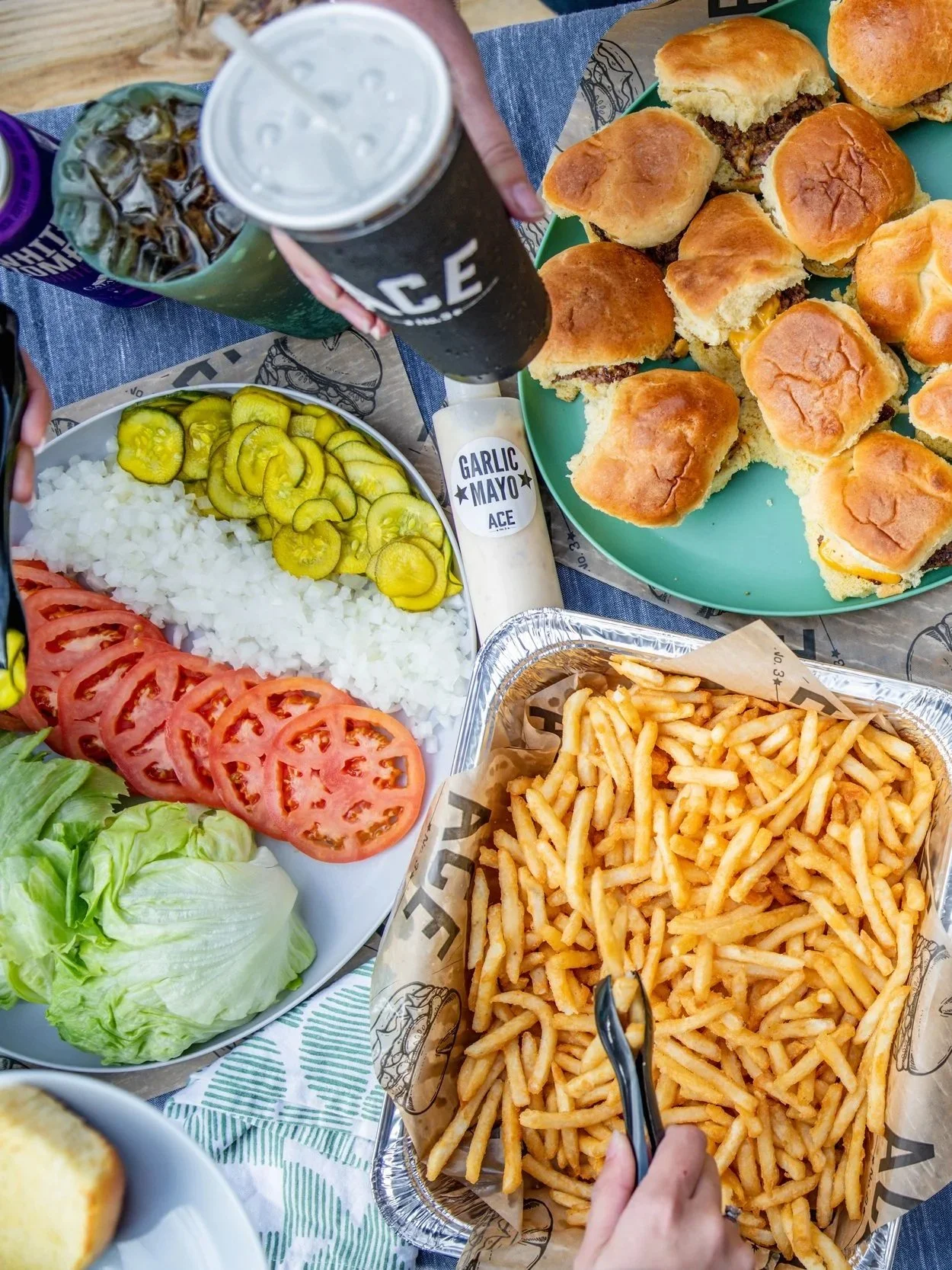 Plate of sliced tomatoes, onions, pickles, and lettuce, along with a tray of French fries, a green plate of cheeseburgers, a cup of soda, and a bottle of garlic mayo sauce on a table.
