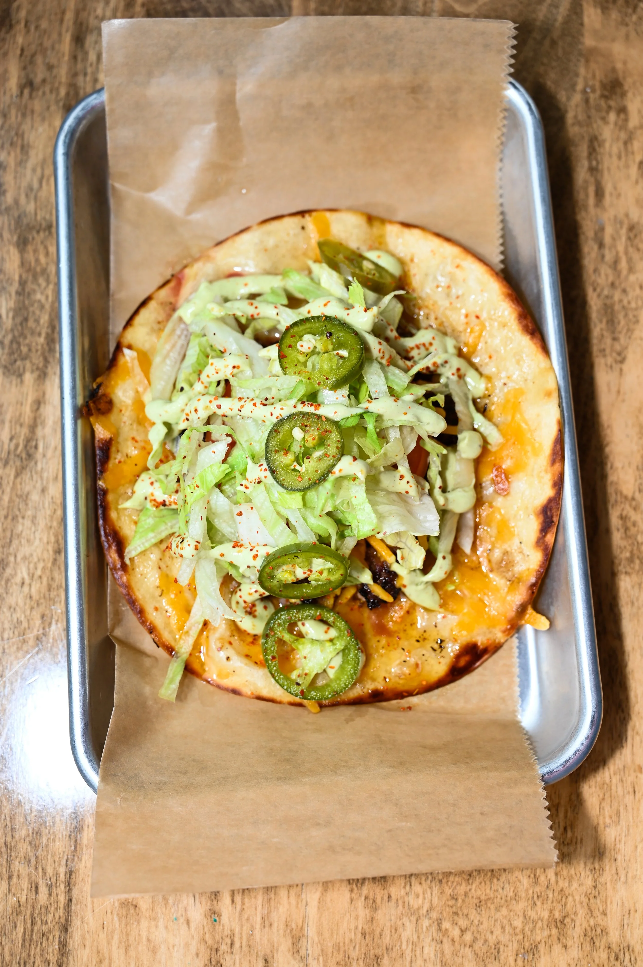 Open-faced pizza topped with shredded lettuce, sliced jalapeños, and a drizzle of sauce, served on a paper-lined tray on a wooden table.