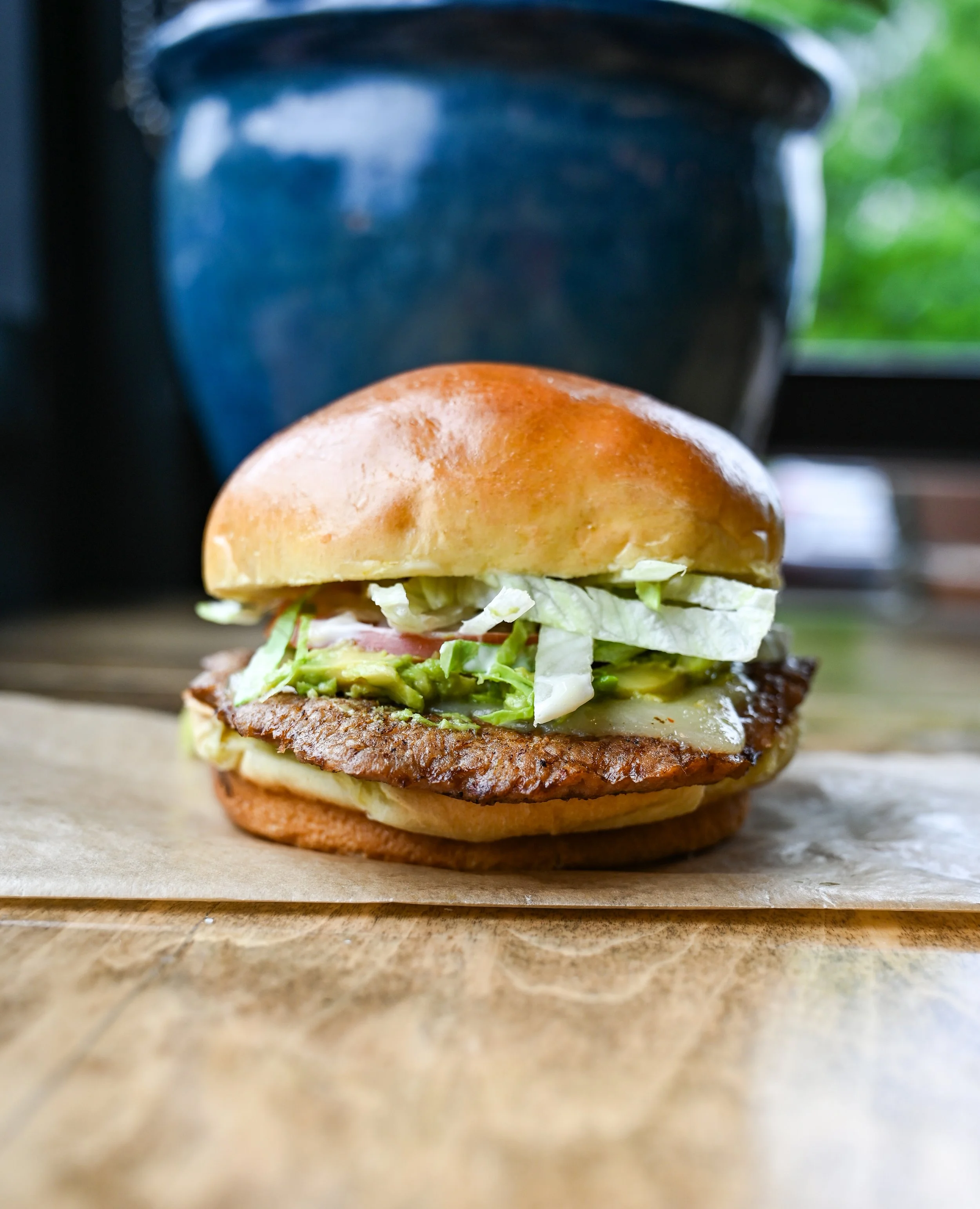 Close-up of a burger with lettuce, tomato, cheese, and a toasted bun, placed on a wooden surface with a blurred background showing a window and green foliage.