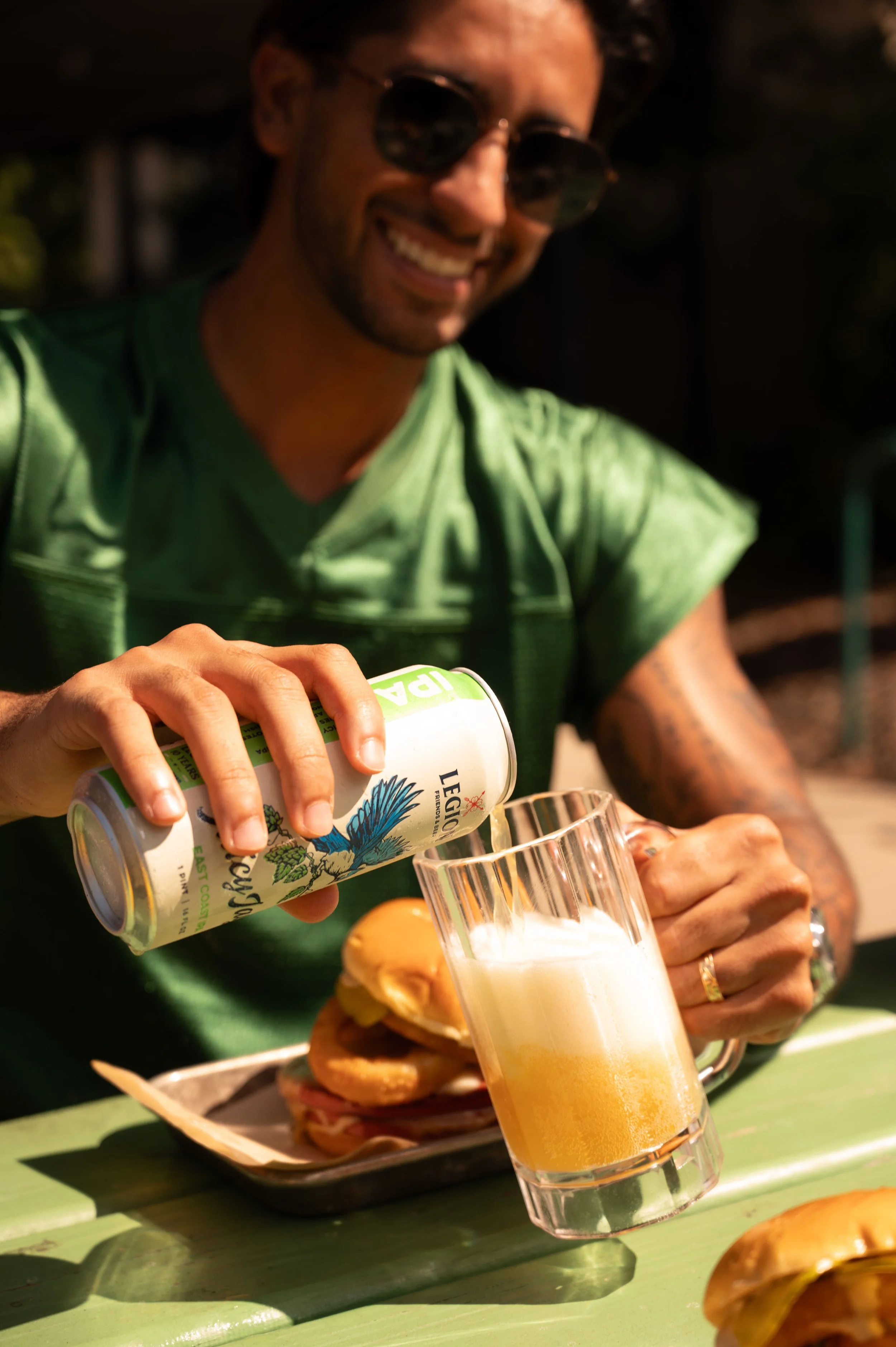A man with a beard, wearing sunglasses and a green shirt, is pouring a beverage into a glass, with a burger plate in front of him, sitting at a green table.