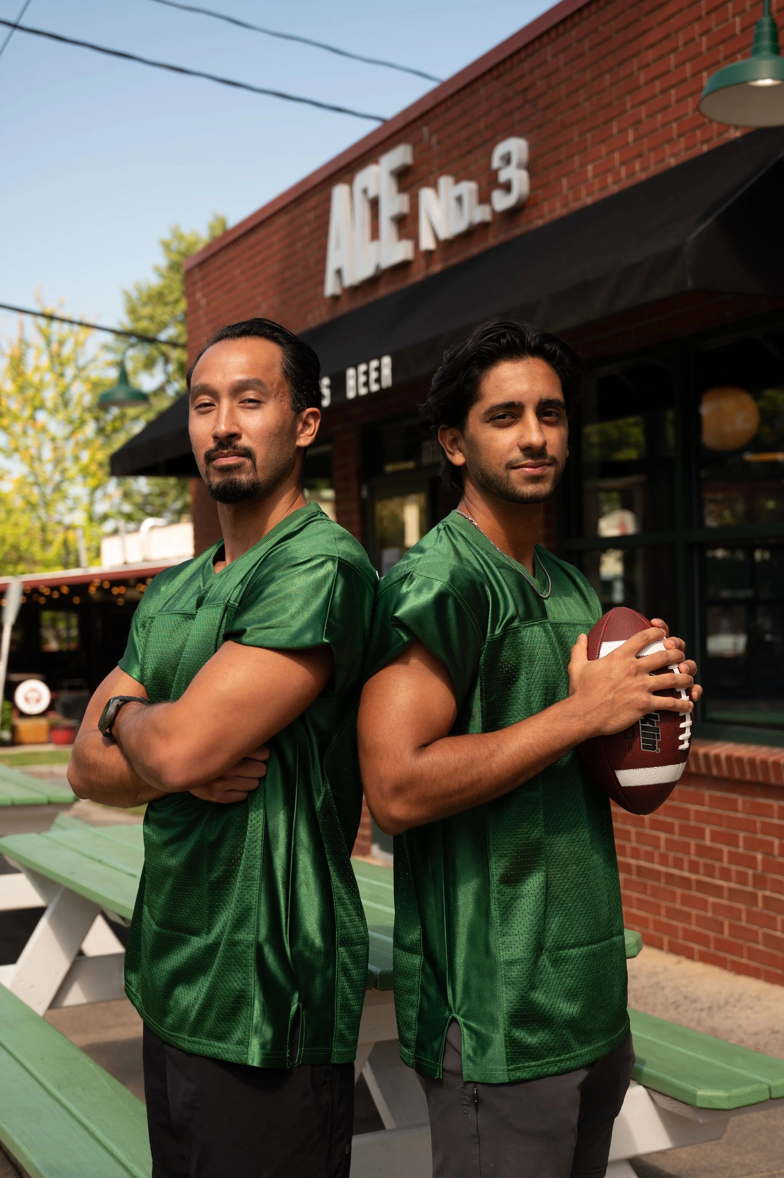 Two men in green football jerseys standing back to back outdoors, one holding a football, in front of a brick building with a sign that says 'Ace No. 3' and some trees in the background.