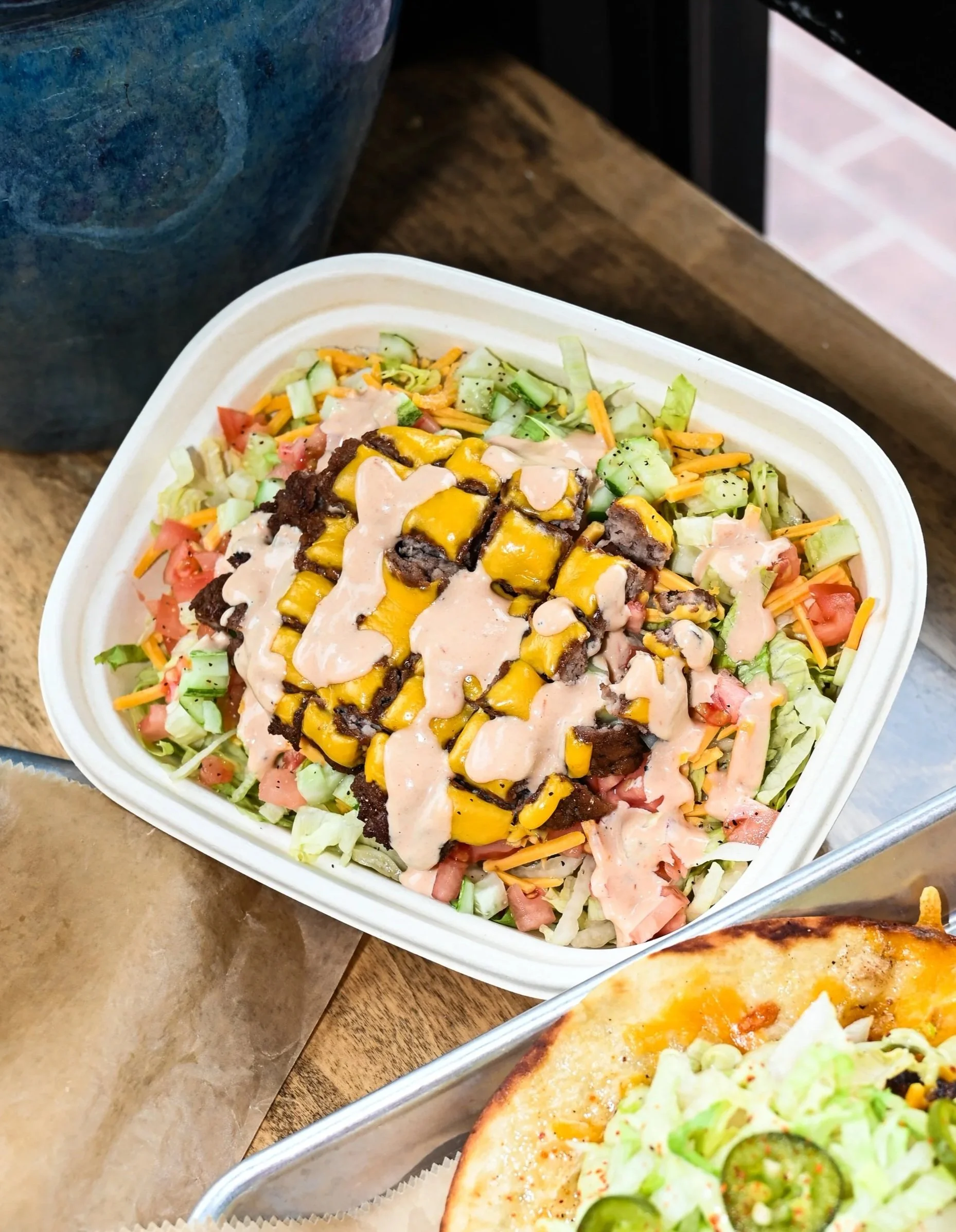 A takeout container filled with chopped lettuce, diced tomatoes, shredded carrots, chopped cucumbers, and chopped ham topped with grilled steak and yellow peppers, drizzled with a creamy dressing. Part of a pizza with lettuce, cheese, and jalapeños is visible in the bottom right corner.