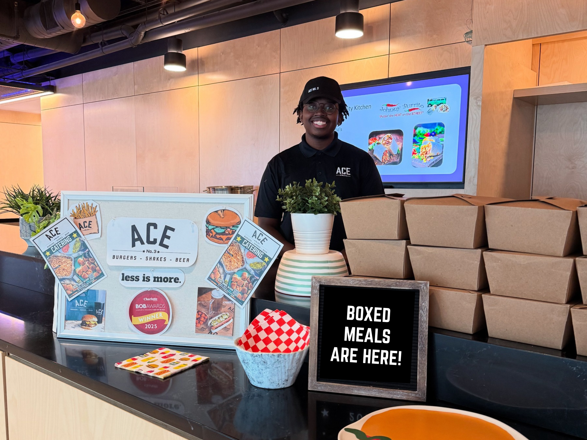 A smiling employee wearing glasses and a black cap, standing behind a counter at a food service station with a digital display and boxed meals stacked to the right. There are promotional signs on the counter for a restaurant called ACE, offering burgers, shakes, and beer, with a plant and decorative items also visible on the counter.