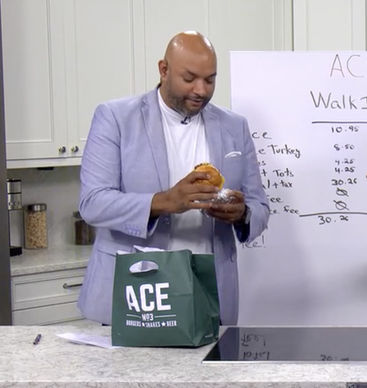 A man in a light gray blazer and white shirt holding a sandwich in a kitchen. There is a green shopping bag with 'ACE' printed on it on the counter, and a whiteboard with a list of prices and items in the background.