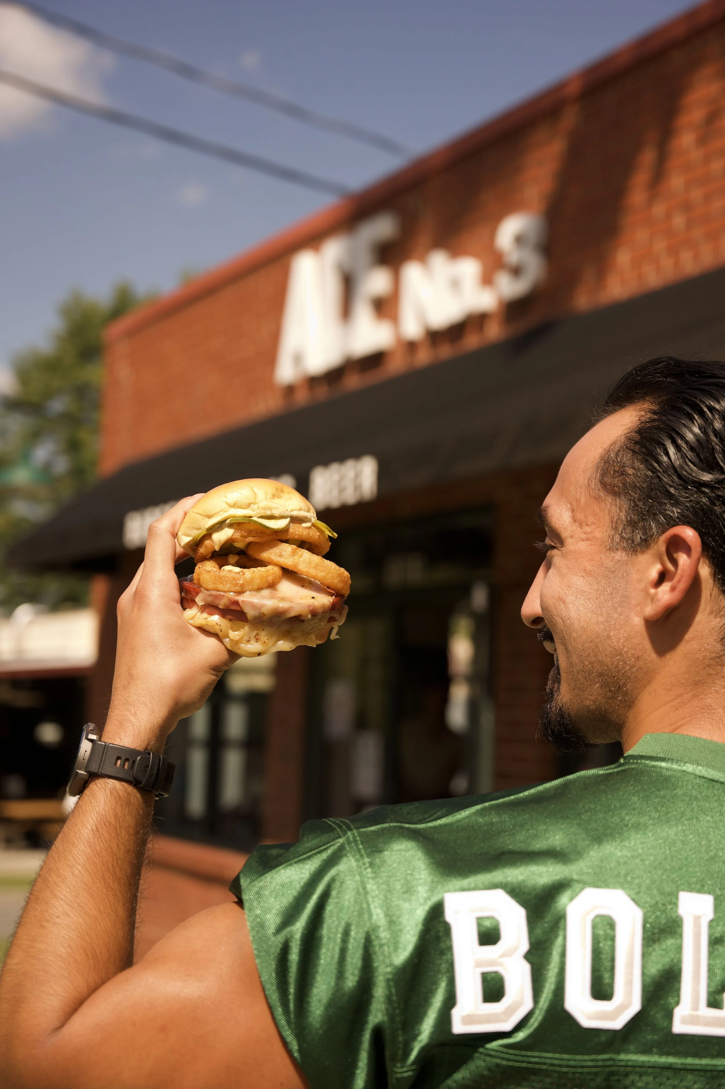 A man holding a fried bologna sandwich in front of a brick building that reads Ace No. 3 in white letters.