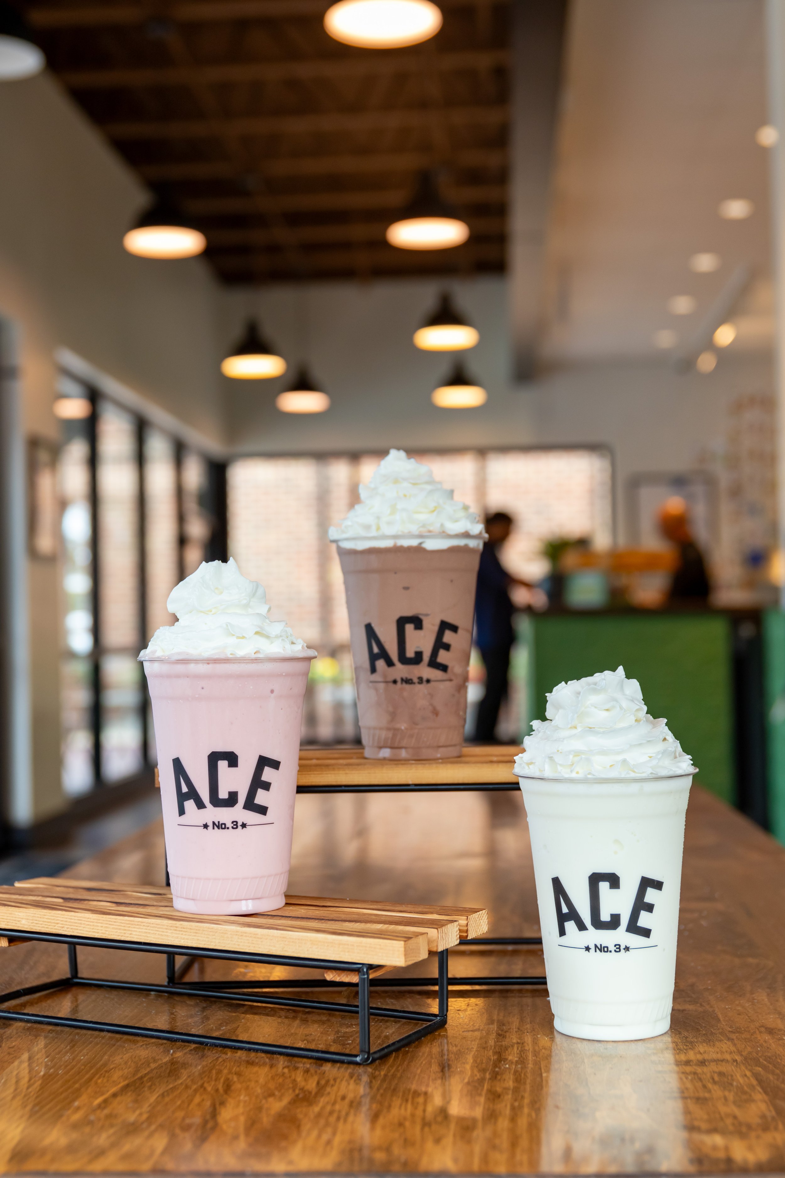 Three beverages with whipped cream on top, served in cups labeled 'ACE No.3', on a wooden table in a cafe-style setting with warm lighting and blurred patrons in the background.