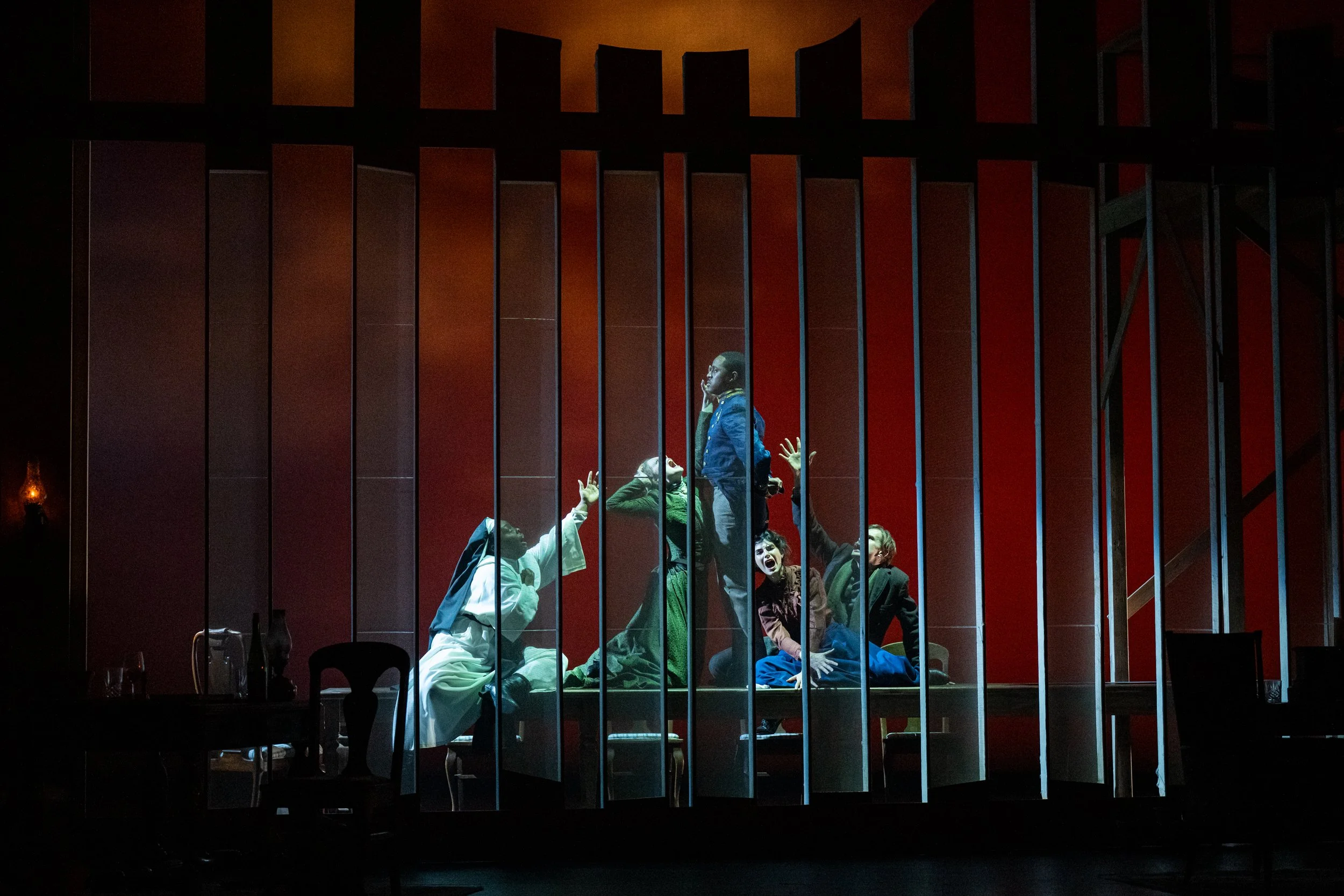 A cast of five pose in a tormented configuration creating a triangle on a table behind a screened set with a flaming red background.