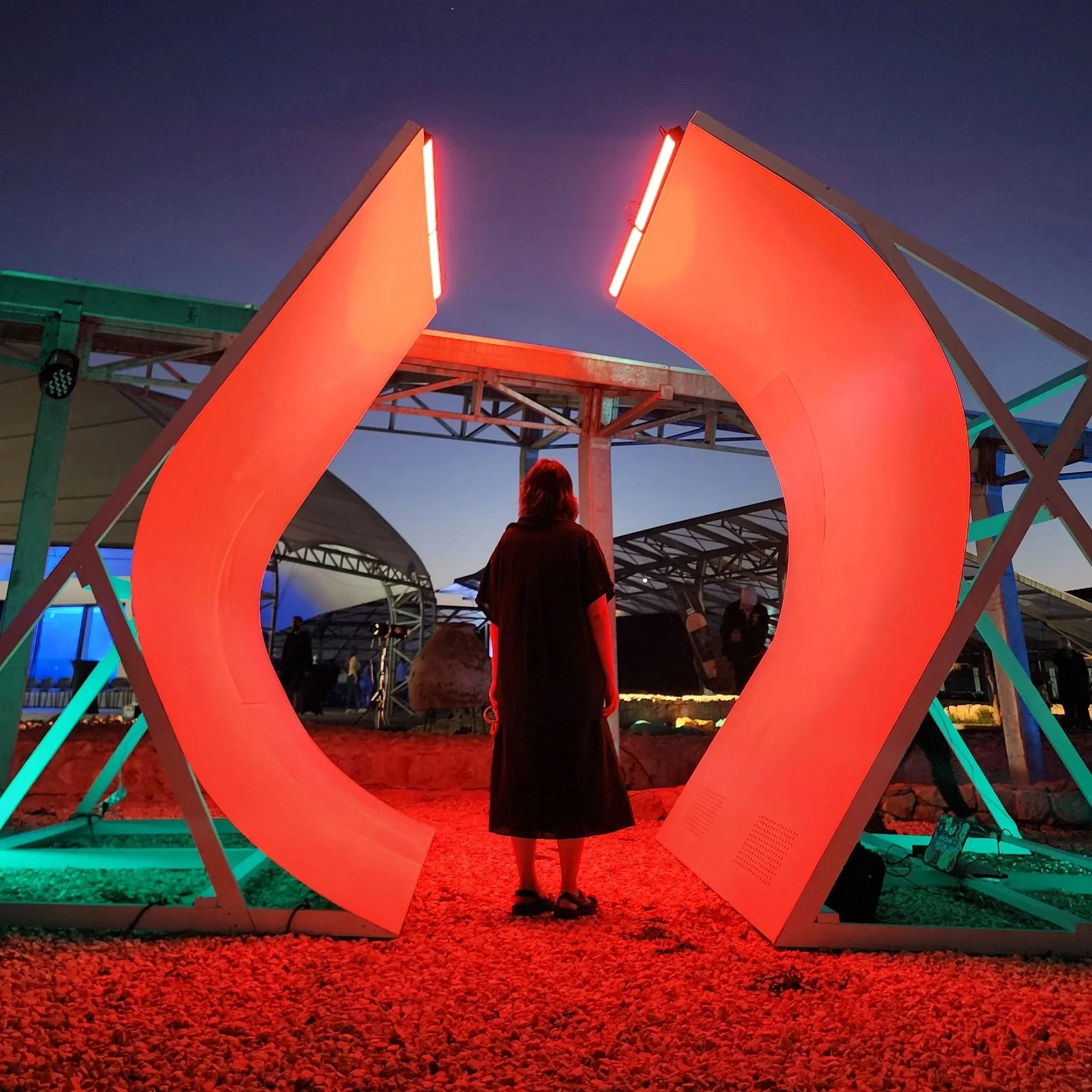 A 4 meter tall sculptural walkway built out of curved plywood is resting on gravel and lit with glowing red-orange light, the outside geometric structure is lit with light teal.