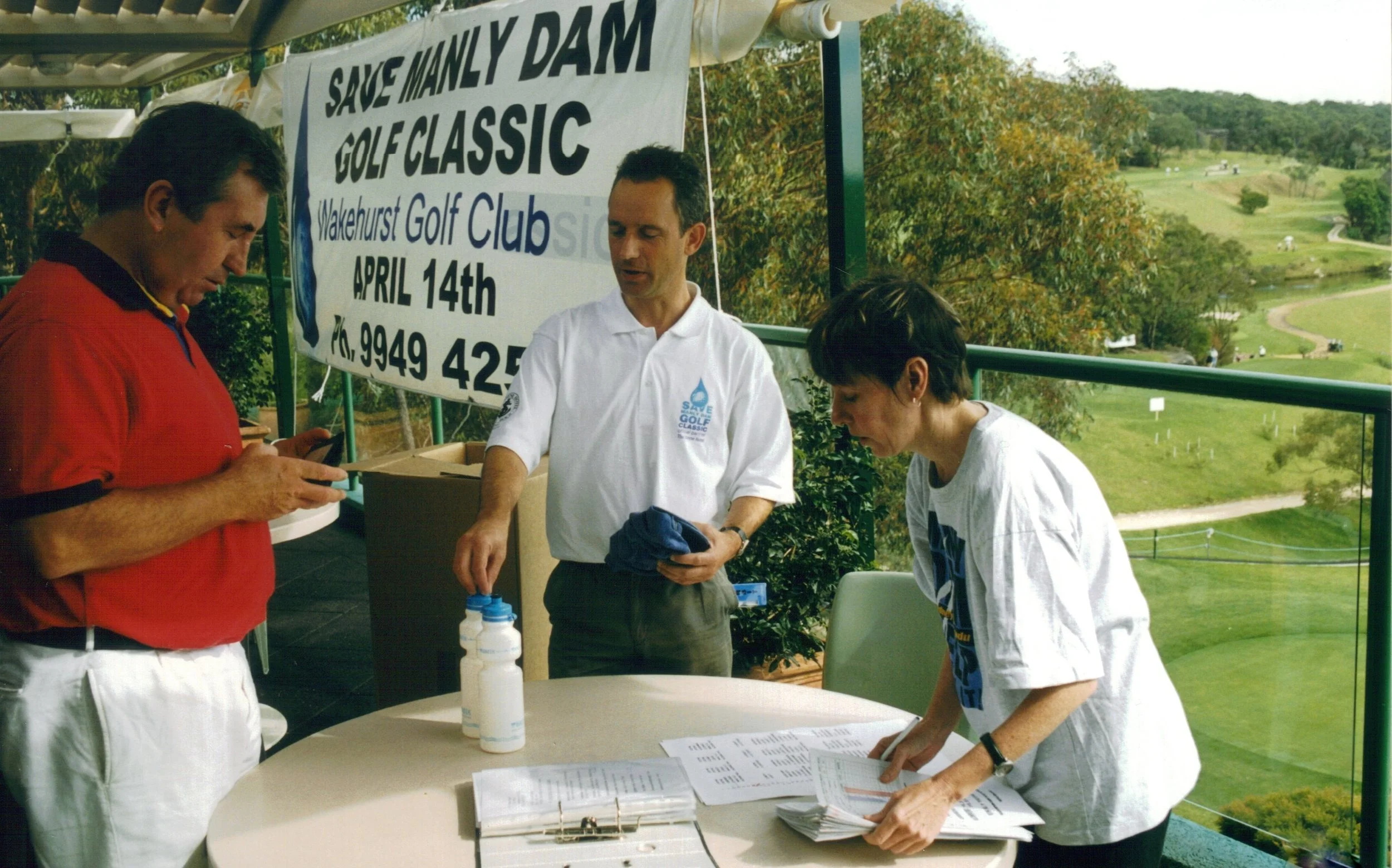 Three people at a table under a tent, preparing for a golf event at Wakehurst Golf Course. A banner behind them reads 'Save Manly Dam Golf Classic April 14th'. The background features a scenic view of a golf course with trees and pathways.