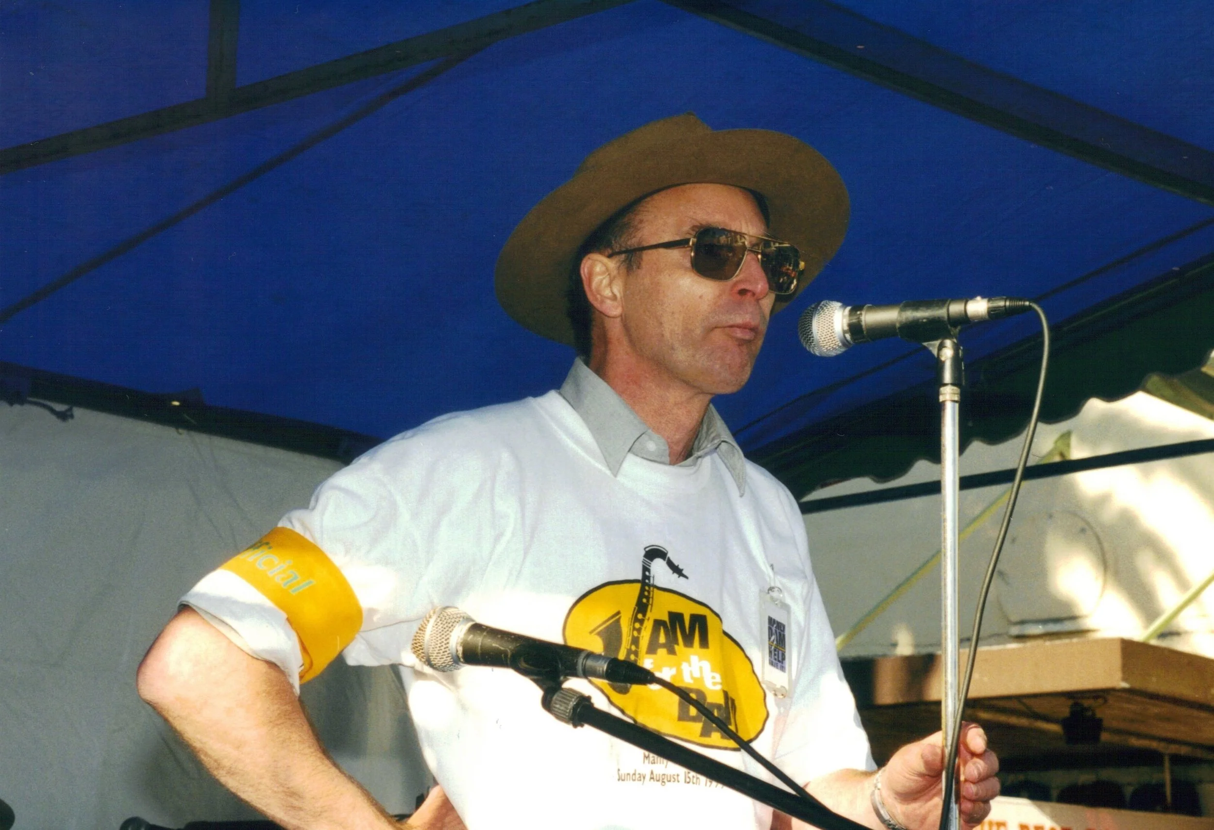 A man wearing sunglasses, a brown hat, and a white t-shirt speaking into a microphone at an outdoor event under a blue canopy.